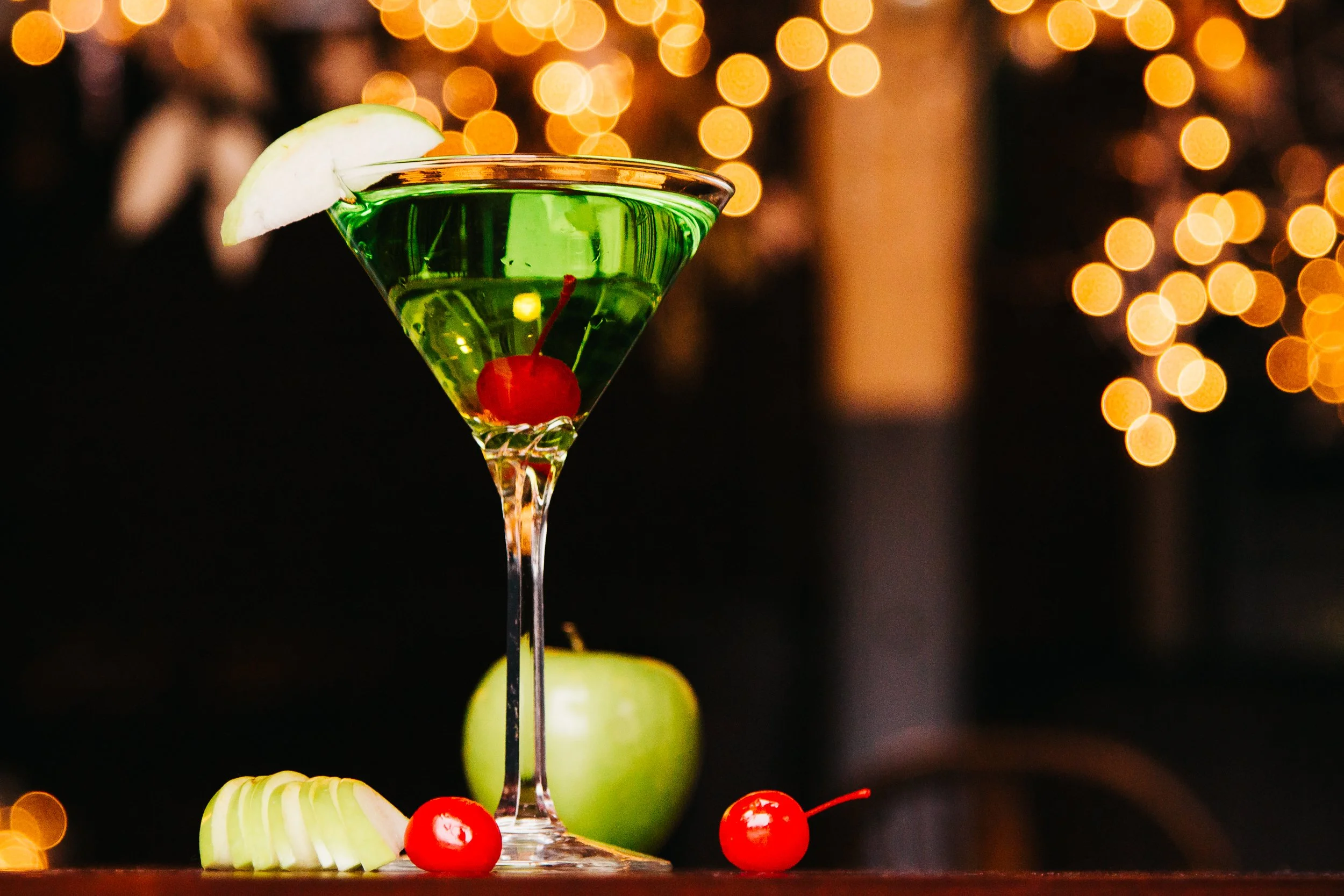 A green cocktail with a cherry and a lemon wedge in a martini glass, with apples, cherries, and apple slices on the table, against a blurred background with warm bokeh lights.