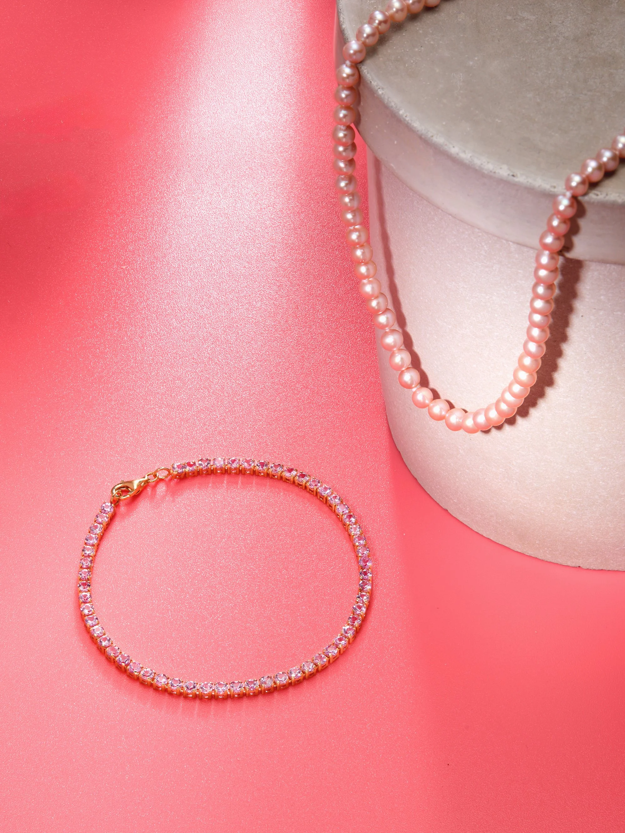 Pink pearl necklace draped over a round jewelry display box and a pink rhinestone bracelet on pink background.