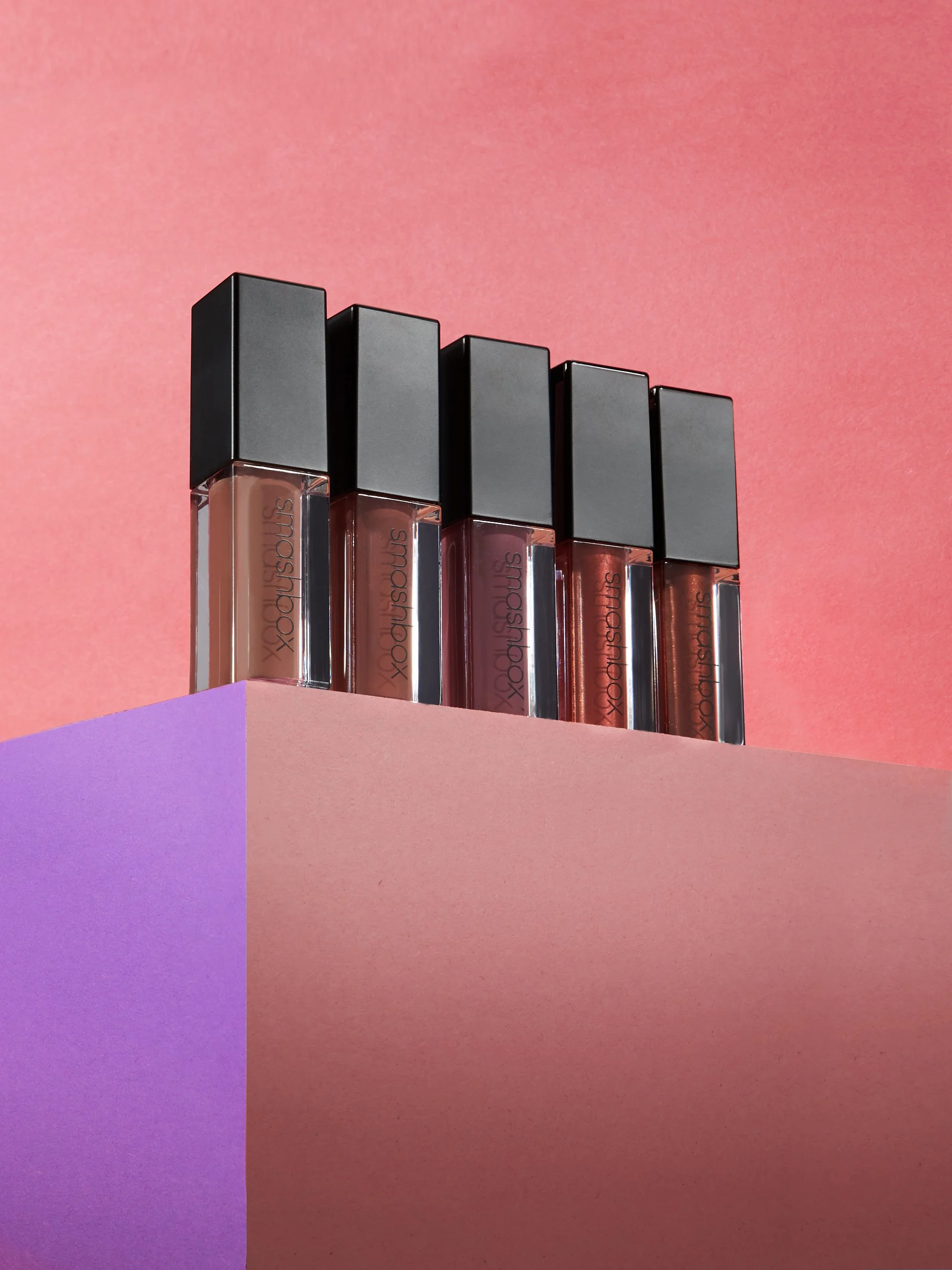 Five lip gloss tubes with black caps displayed on a pink and purple surface against a pink background.