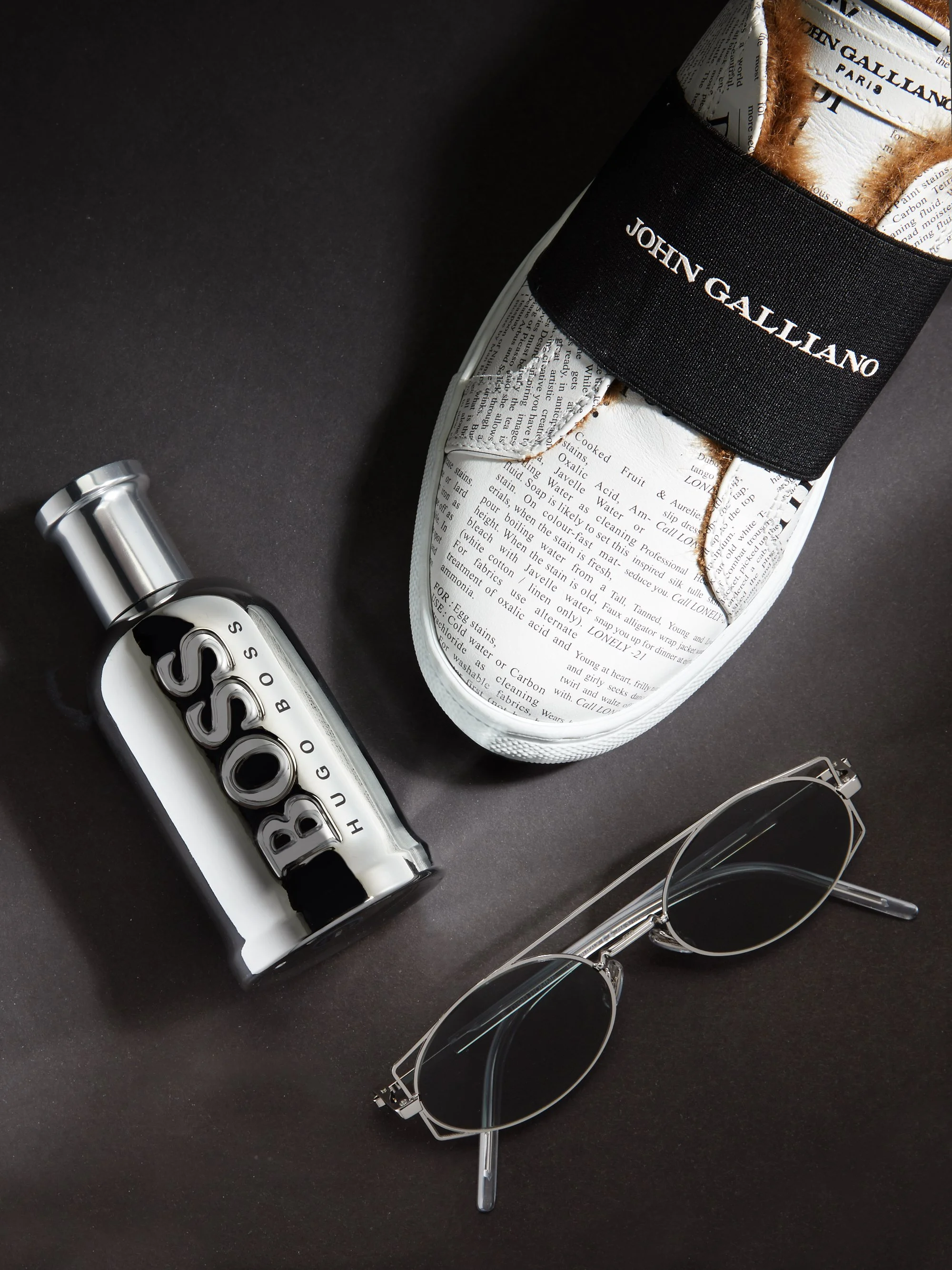 A pair of white sneakers with newspaper print design and black elastic band labeled 'JOHN GALLIANO' around the ankle, a black and white Hugo Boss cologne bottle, and a pair of wire-framed sunglasses with dark lenses arranged on a black surface.
