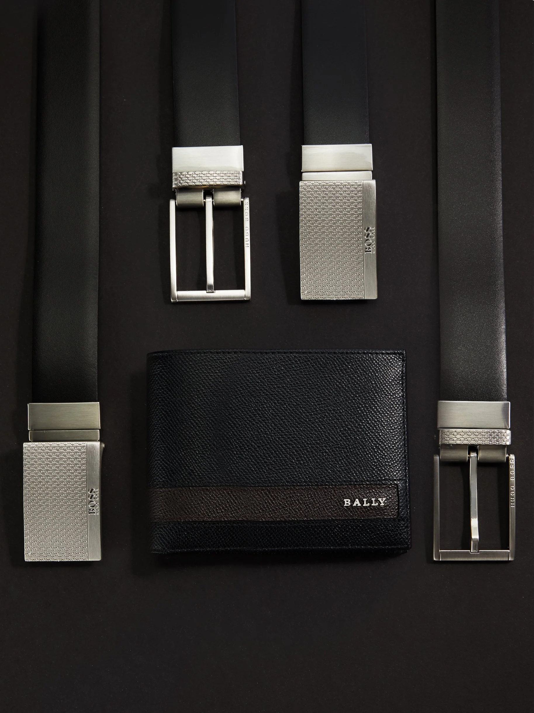 A collection of men's fashion accessories on a black surface, including two black belts with silver buckles, a black textured wallet with a brown stripe and «BALLY» branding, and two silver card holders with «BOSS» branding.