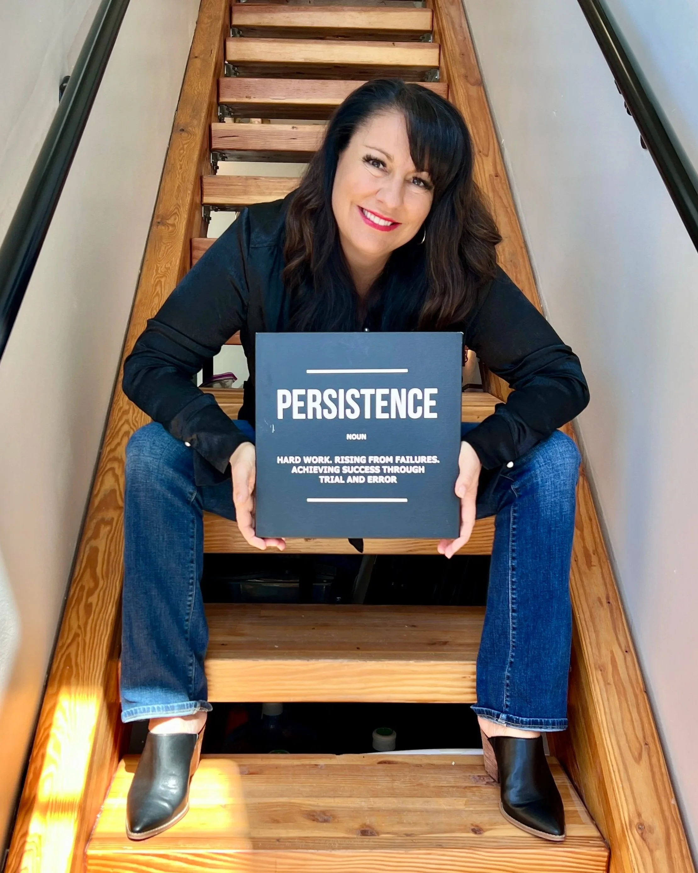 Woman sitting on wooden stairs holding a book titled "Persistence". She is smiling, wearing a black top, blue jeans, and black boots.