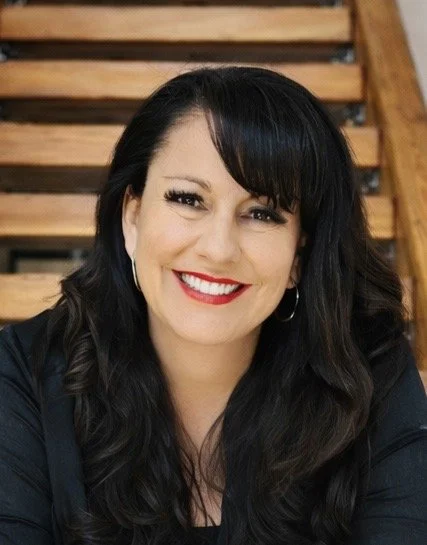 A woman with long black hair and red lipstick smiling in front of a wooden staircase.
