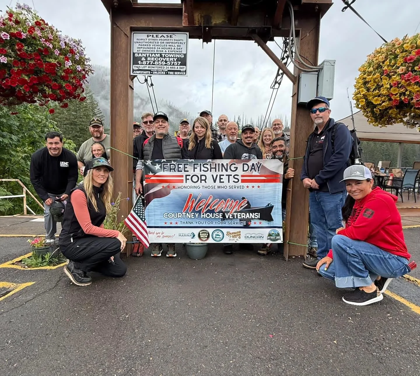 Group of people holding a banner that reads 'Free Fishing Day for Vets' at a restaurant or lodge. The banner also says 'honoring those who served' and welcomes courthouse veterans. The people are smiling, some wearing casual clothing, hats, and sunglasses. There are flowers and hanging baskets on either side of the group, and a parking lot in the background.