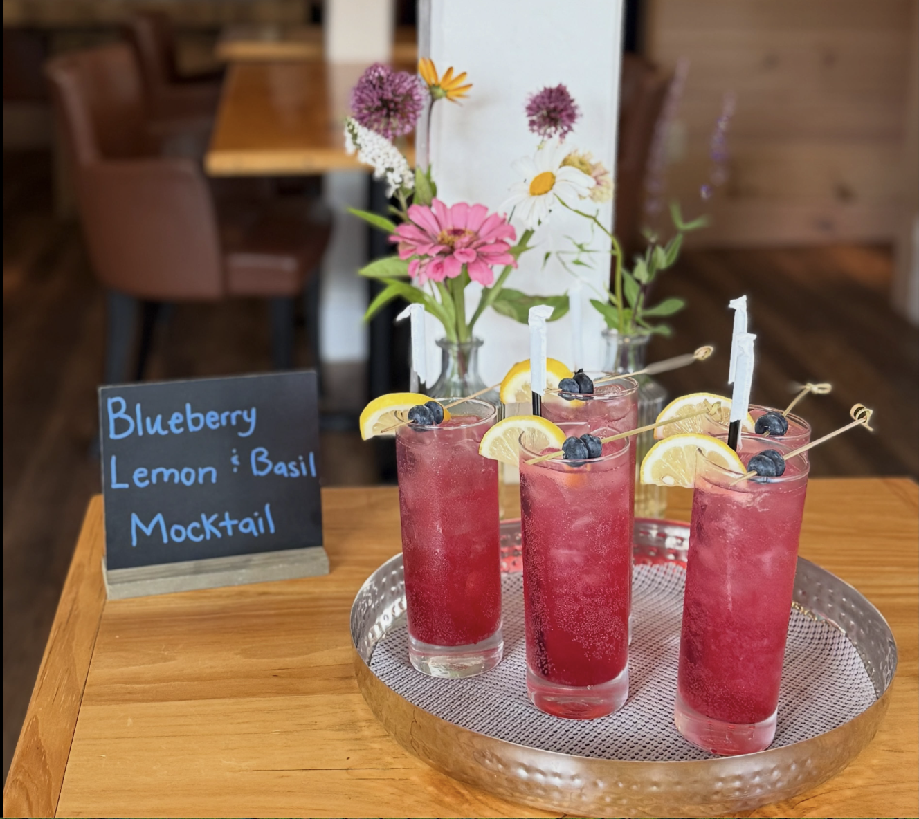 5 tall glasses containing blueberry, lemon, basil mocktails garnished with a lemon wedge and blueberries on a silver tray