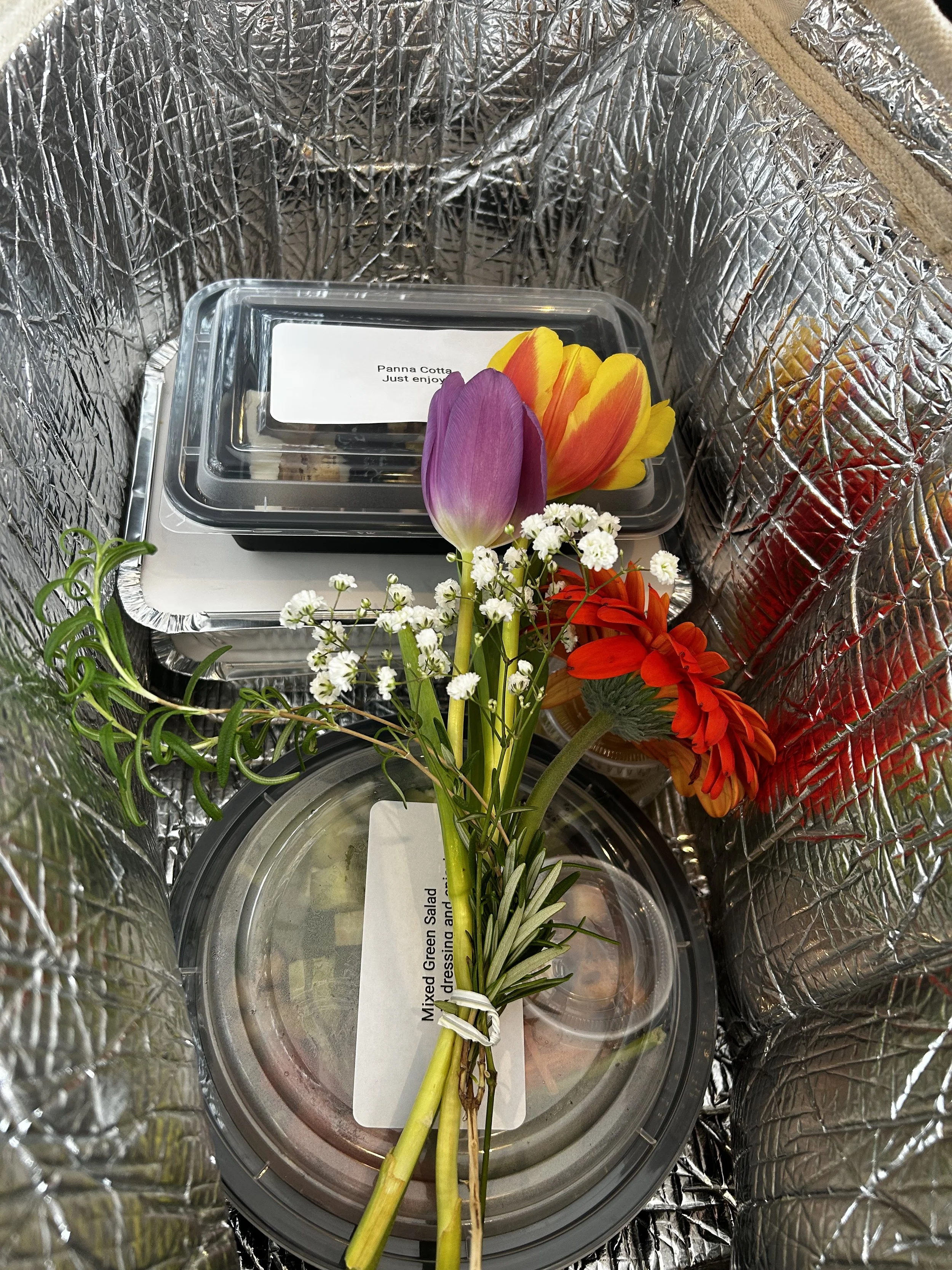 Inside of a cooler bag sits a freshly prepared meal topped with a floral bouquet
