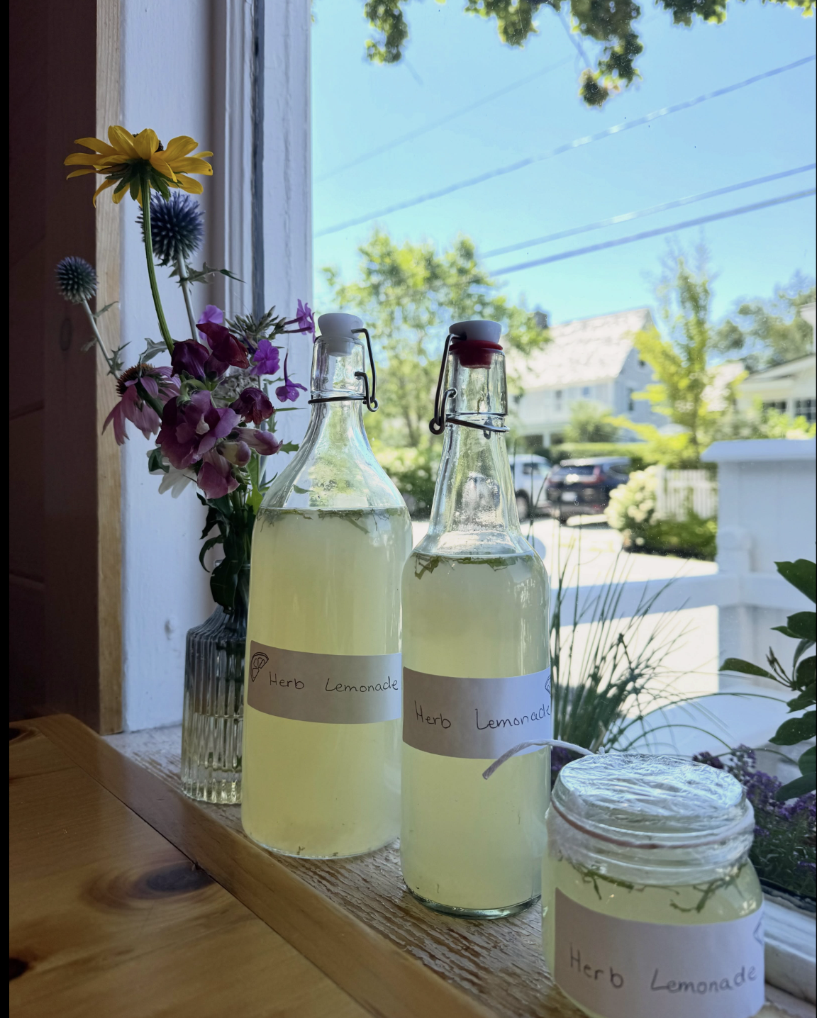 A windowsill with a small bouquet of wildflowers, two tall glass jars, and one squat jar filled with herb lemonade