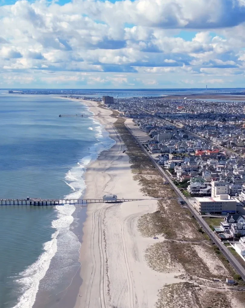 Aerial view of a beach with sandy shoreline, ocean waves, piers extending into the water, and a densely populated coastal town with houses, buildings, and infrastructure under a partly cloudy sky.