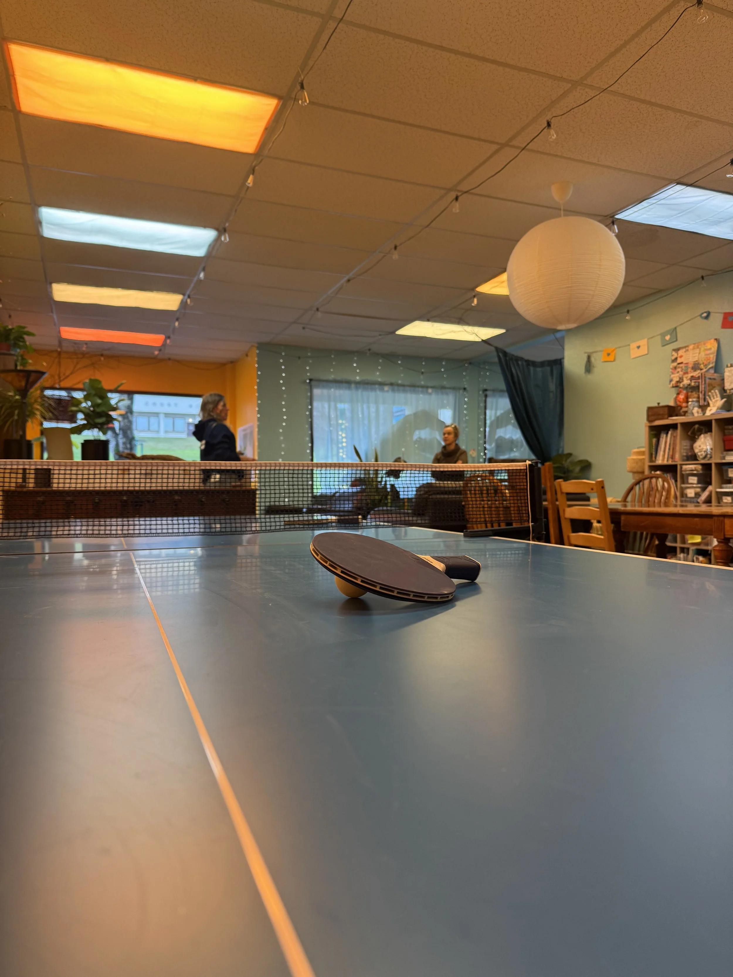 Two women, named Kika and Dana, stand in the background as a low shot of a ping-pong paddle rests on its ball atop a ping-pong table.