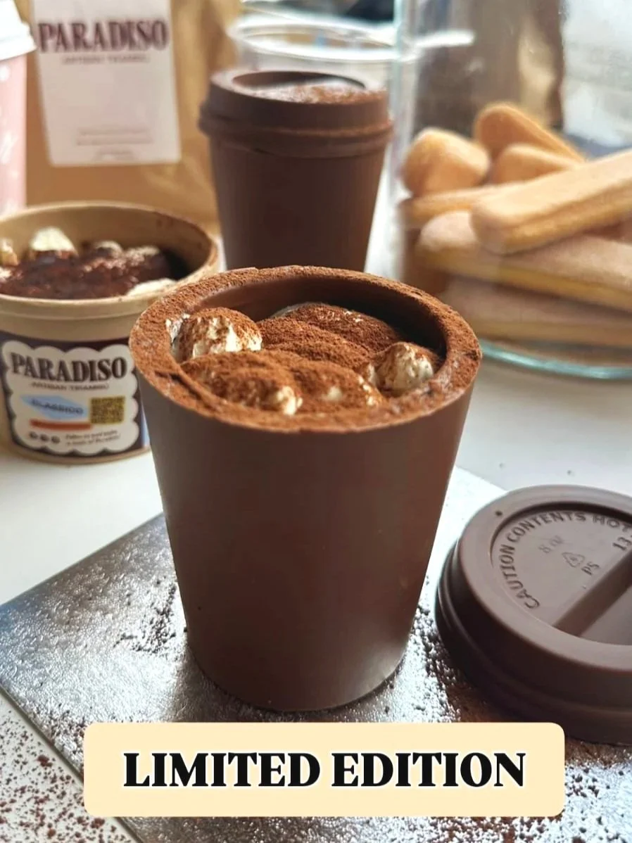 A limited edition edible chocolate cup topped with tiramisu, marshmallows, with a coffee cup, spoon, and savoiardi in the background.