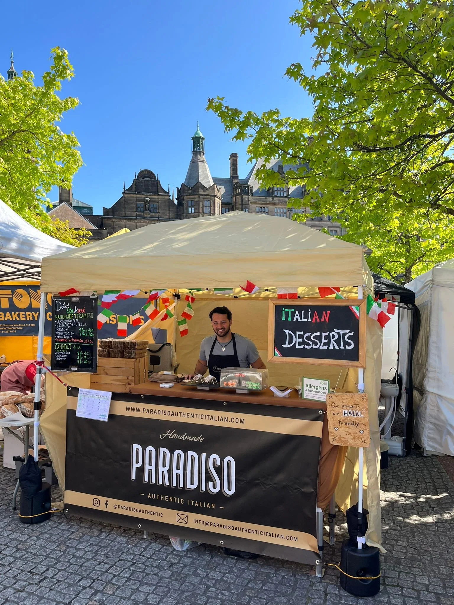 Outdoor market stall selling Italian desserts with a vendor standing behind a table, decorated with Italian flags, and signage displaying "Italian Desserts." Trees and a historic building are visible in the background.