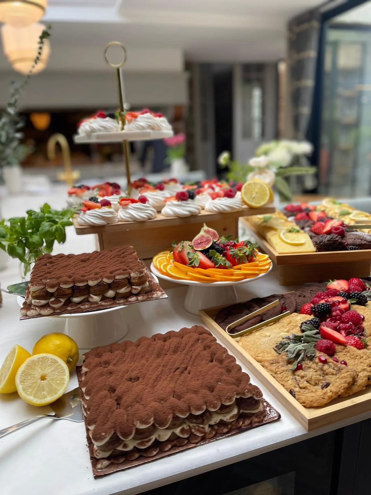A table with various desserts, including tiramisu, pavlova topped with berries, cookies, sliced fruits, and lemons.
