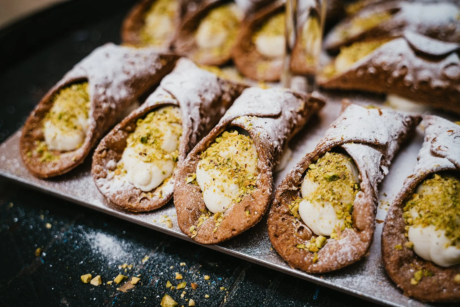 Cannoli with ricotta filling, topped with pistachio crumbs and powdered sugar on a tray.