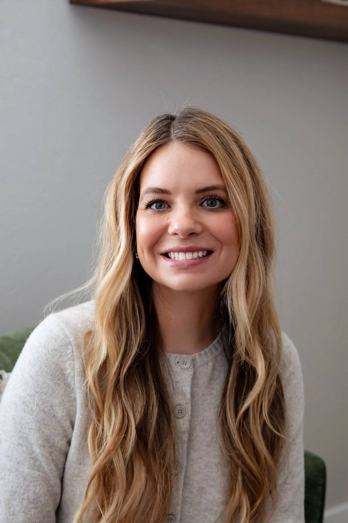 A woman with long blonde hair smiling, wearing a white button-up shirt, sitting indoors with a lamp in the background.