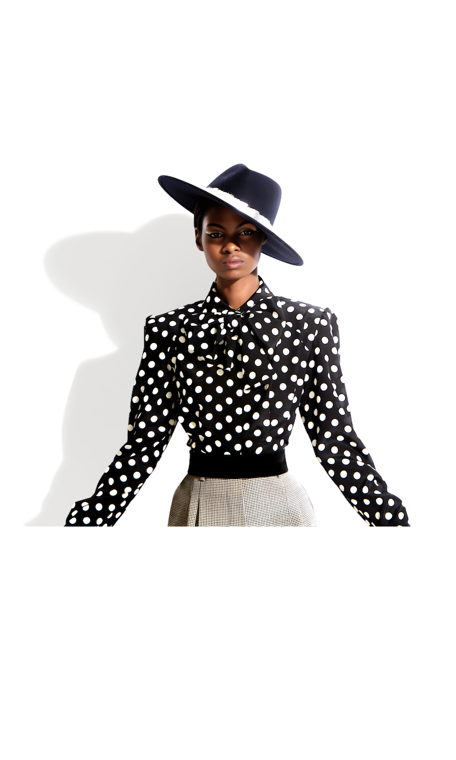 A woman wearing a black wide-brimmed hat with white accent, a black polka dot blouse with bow tie, and beige high-waisted pants, standing against a white background.