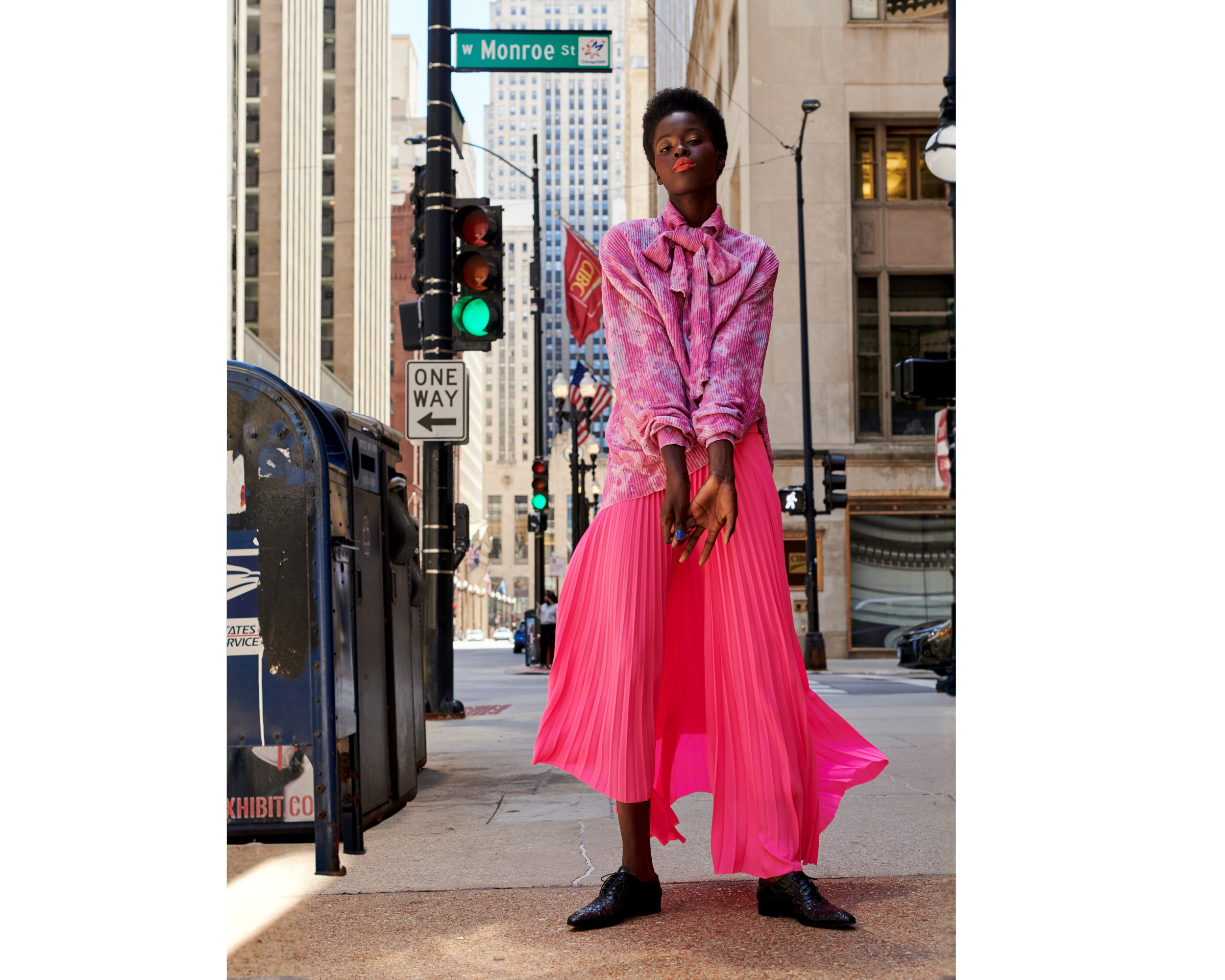 A woman in a pink pleated skirt and pink blouse with a bow stands on a city sidewalk, with tall buildings and street signs including W Monroe Street visible in the background.
