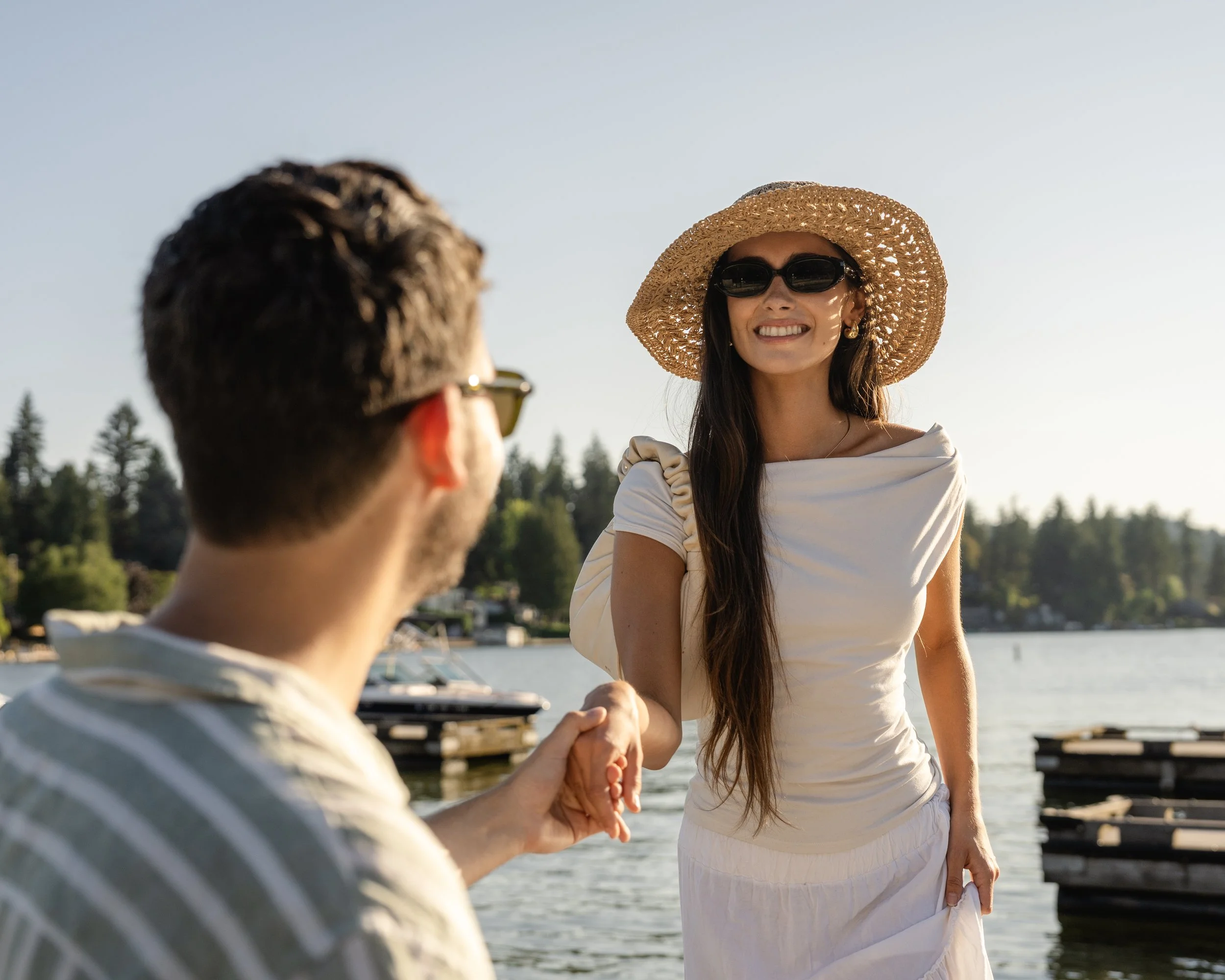 A woman wearing a straw hat, sunglasses, and a white dress smiling while holding hands with a man near a lake, with boats and trees in the background on a sunny day.
