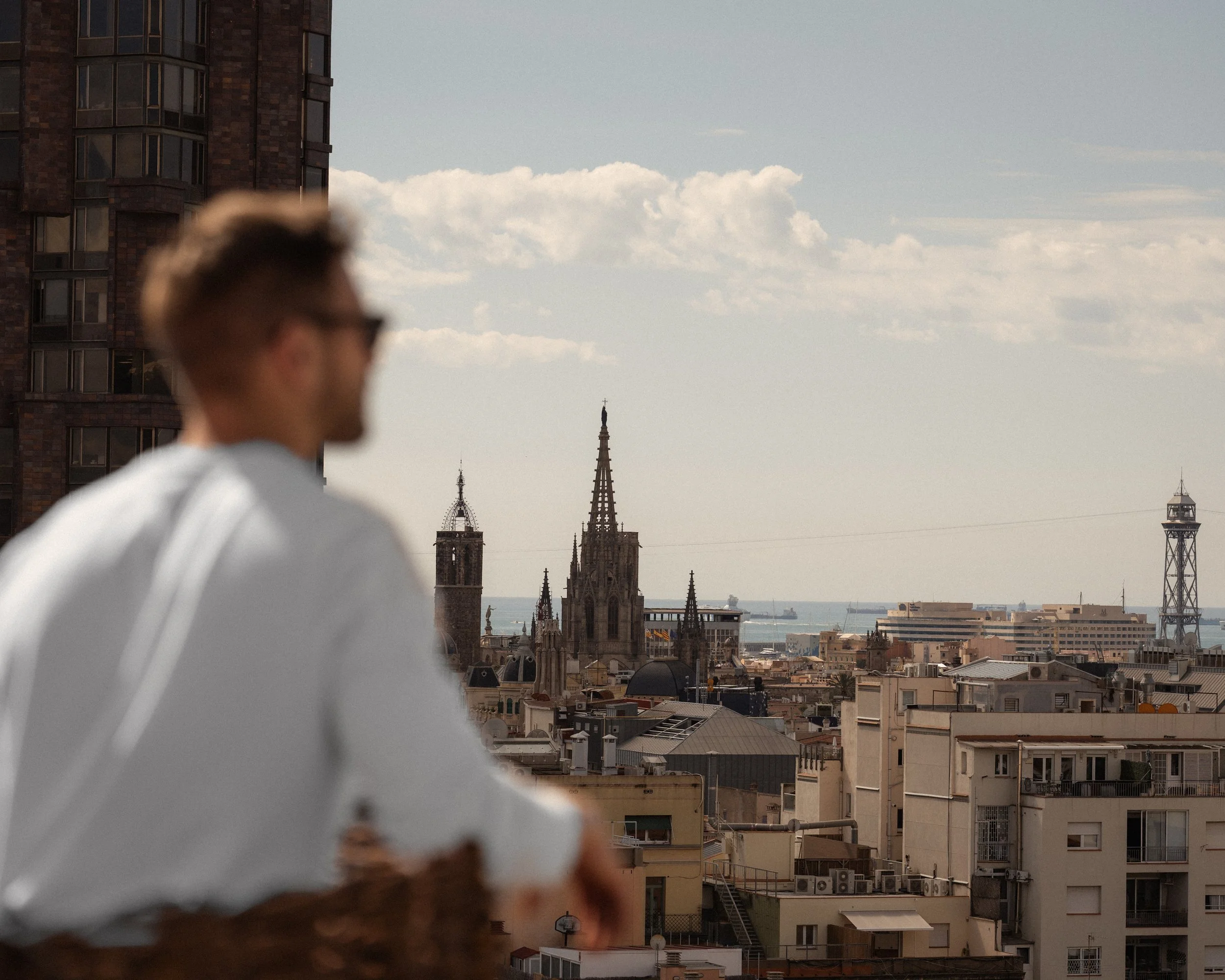 Blurred man with sunglasses and white shirt in foreground, cityscape with historic buildings and church steeples, and waterfront in the background.