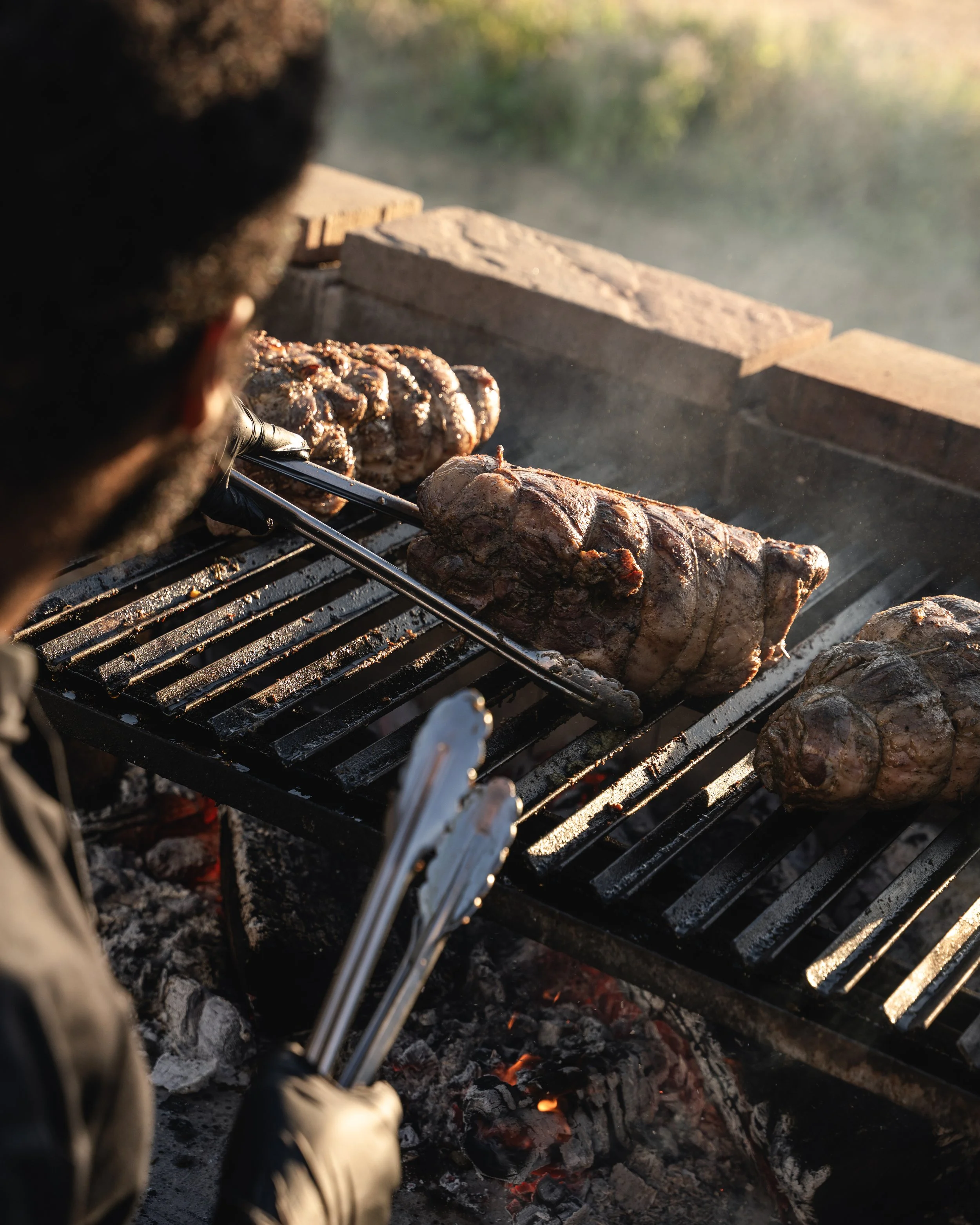 Person grilling large cuts of meat, including steaks and sausages, over an open flame outdoors.