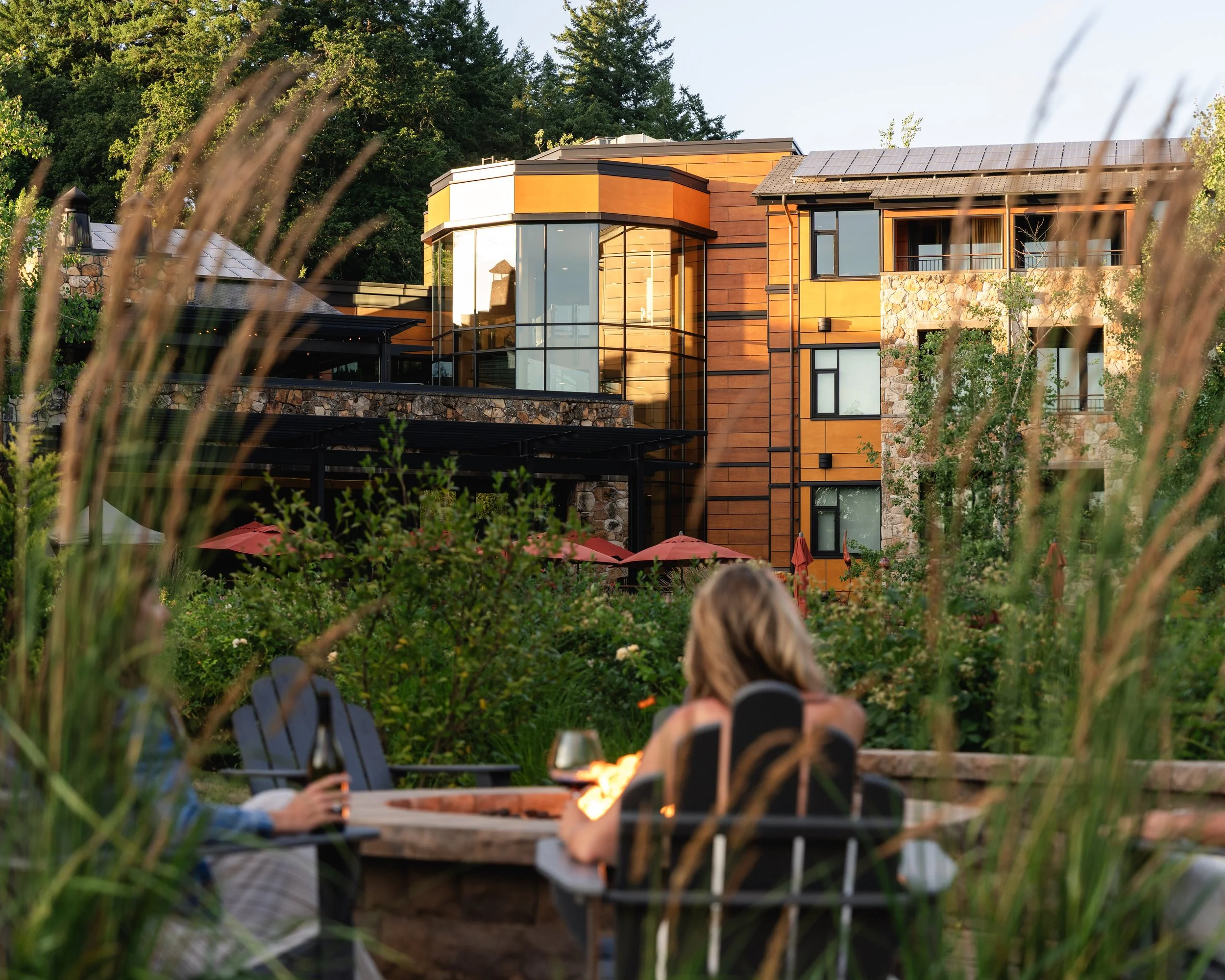 People sitting around a fire pit in a garden with a modern multi-story house in the background, during sunset.