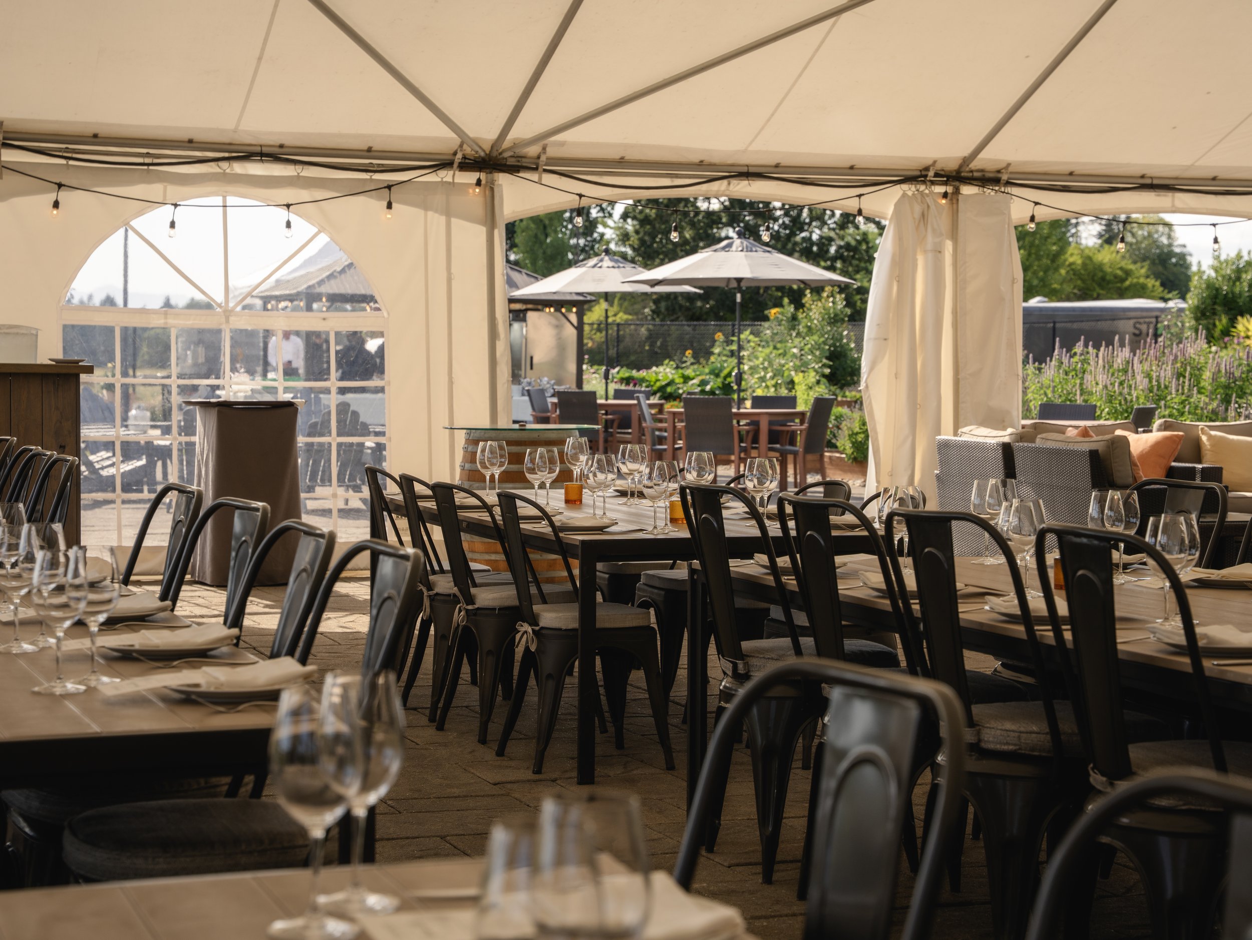 Empty outdoor dining area with tables, chairs, and wine glasses under a large white tent, with patio furniture and umbrellas outside.