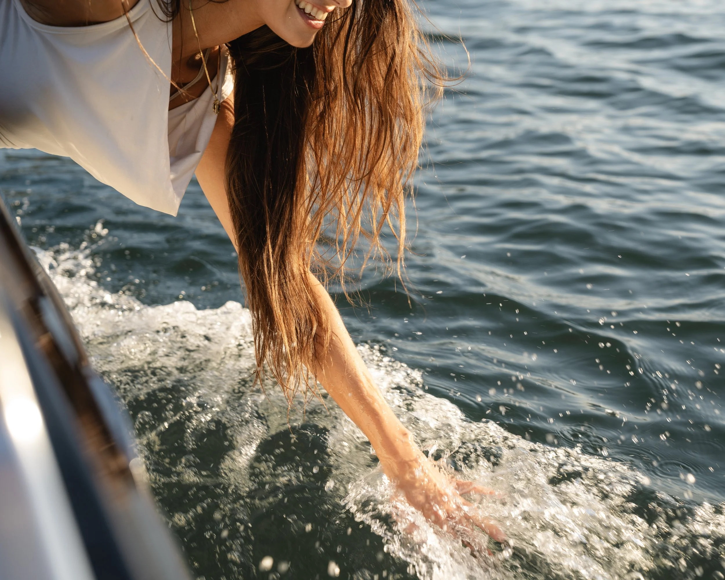 A woman with long red hair reaching into water from a boat, smiling, wearing a white shirt.