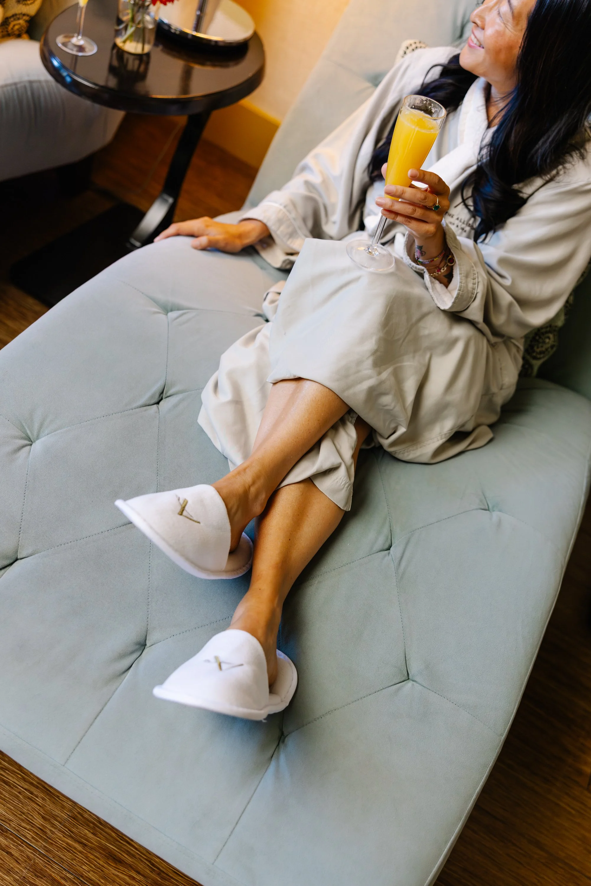 A woman sitting on a light gray tufted sofa, wearing a beige robe and white slippers, holding a glass of orange juice, smiling, in a cozy living room.