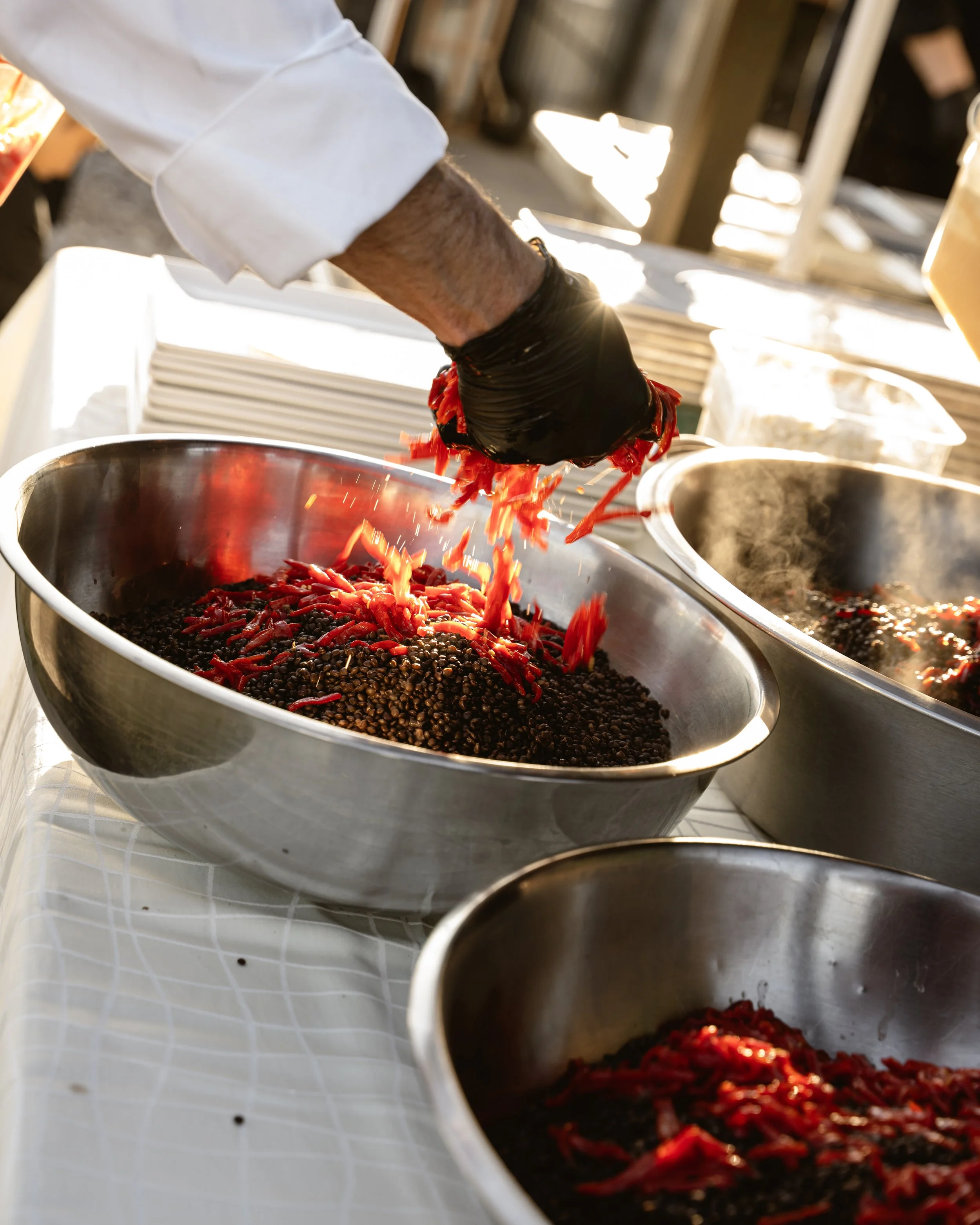 Person wearing black gloves and a white long sleeve shirt adding red shredded peppers into black beans in a large stainless steel bowl.