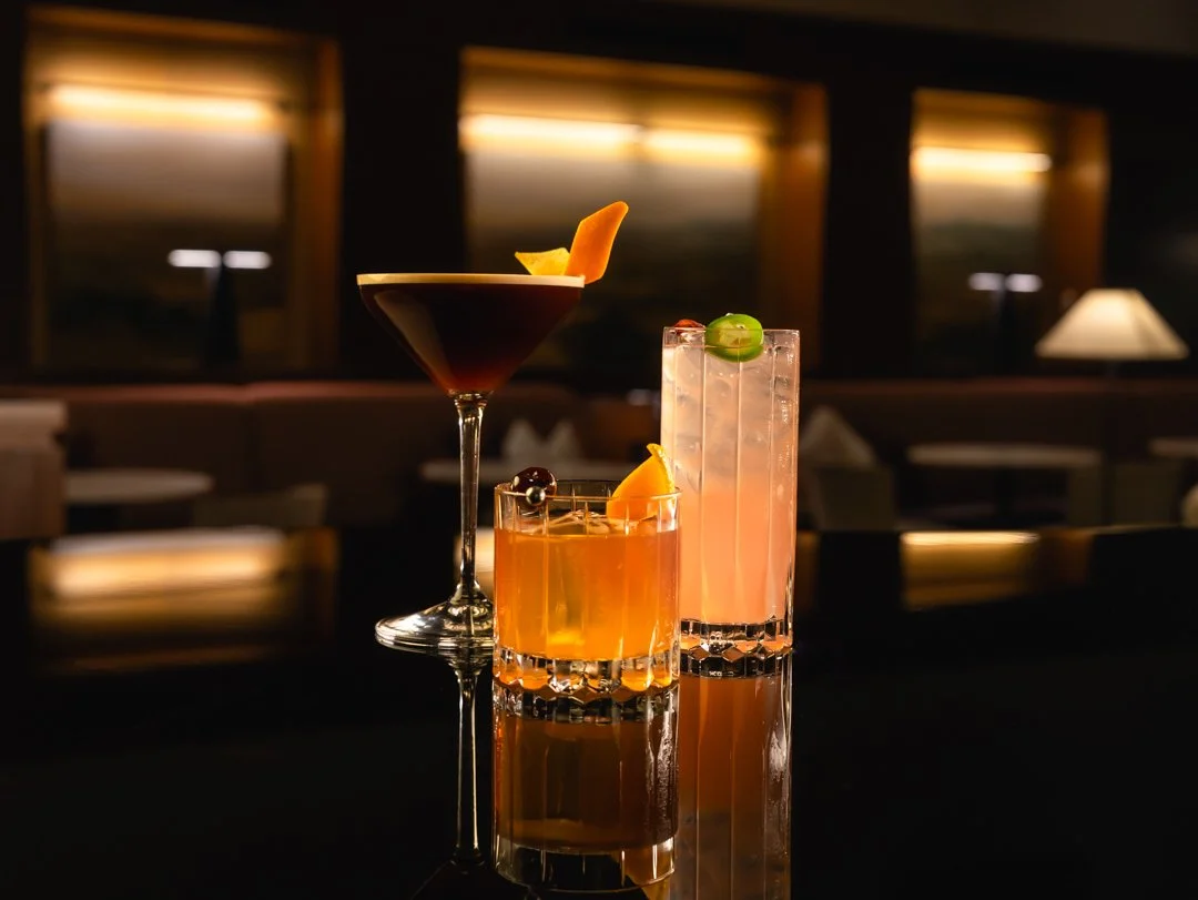 Three colorful cocktails on a reflective surface in a dimly lit bar or lounge, with a dark background and warm lighting.