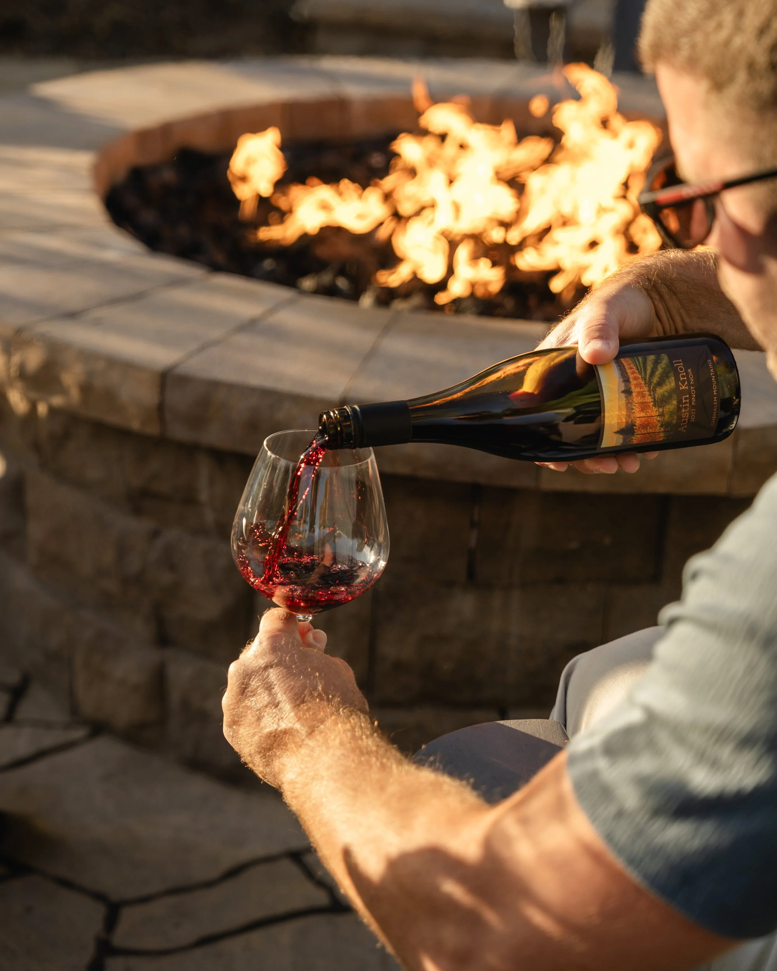 A man wearing sunglasses is pouring red wine from a bottle into a wine glass near a lit outdoor fire pit.