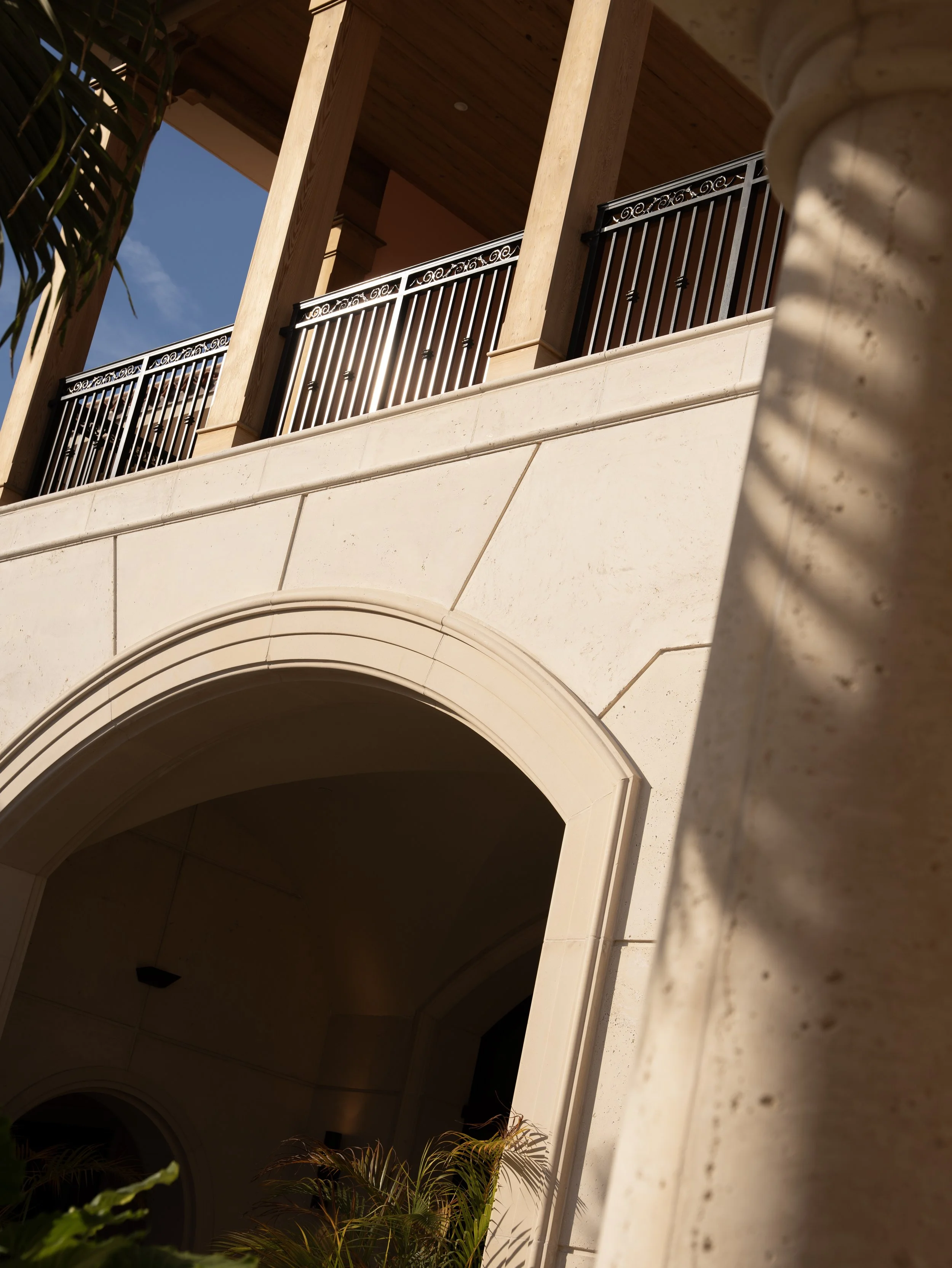 A view of the exterior of a building showing a large archway at the entrance, with a balcony above it featuring decorative black metal railings, and tall beige columns supporting the balcony. Sunlight and shadows are visible, with some greenery at the bottom.