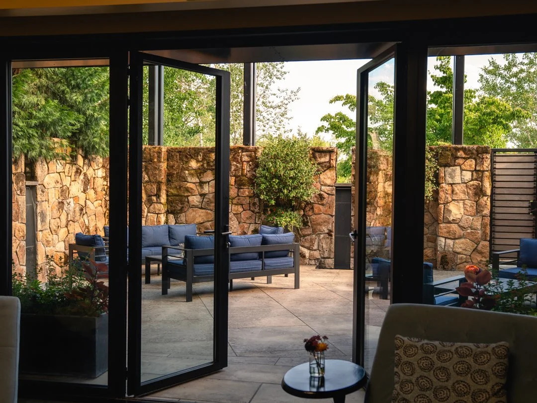 View of an outdoor patio with stone walls, blue outdoor sofas, and greenery, seen through open glass doors from inside a building.