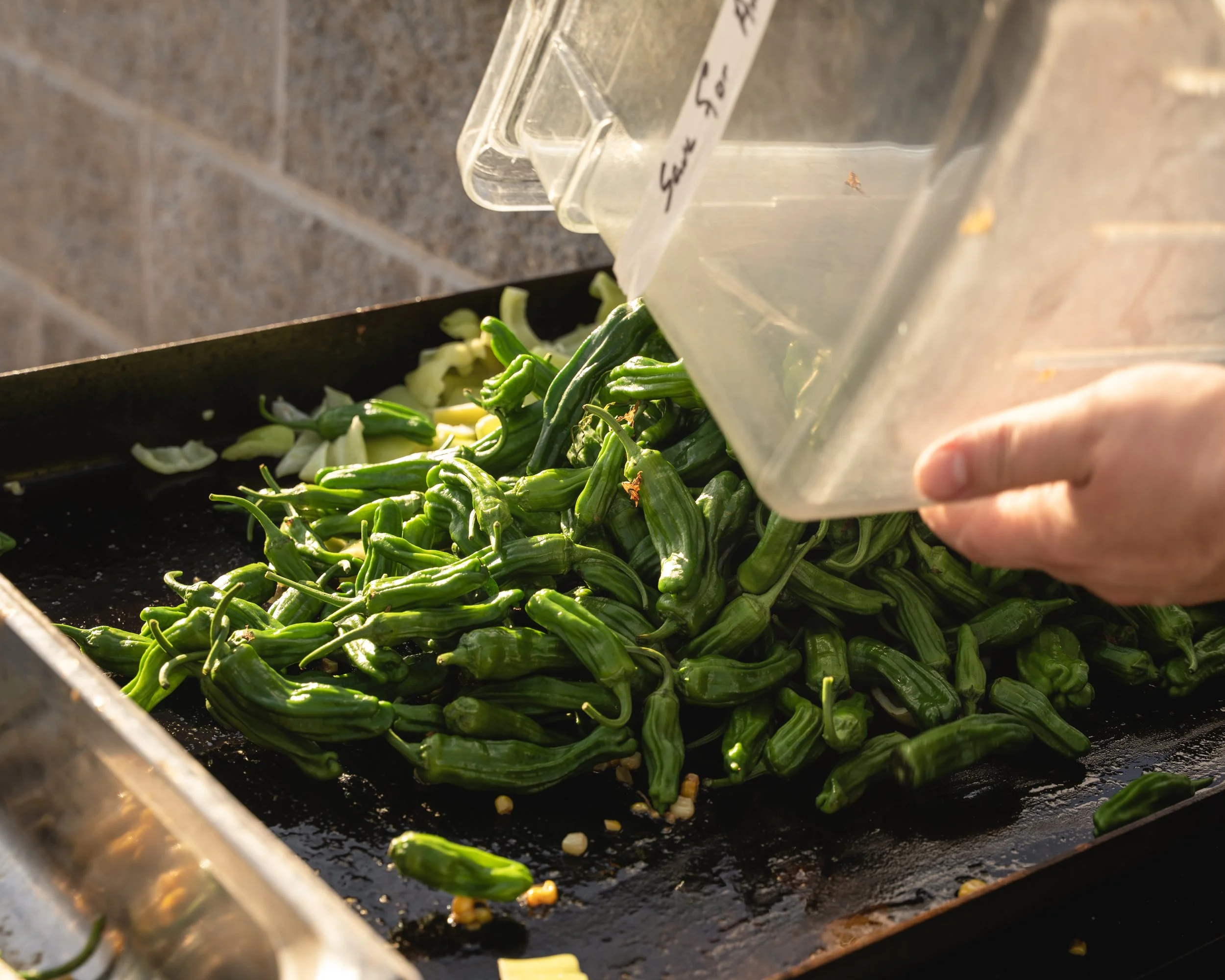 Person pouring sliced green peppers onto a black cooking surface with chopped garlic in the background.