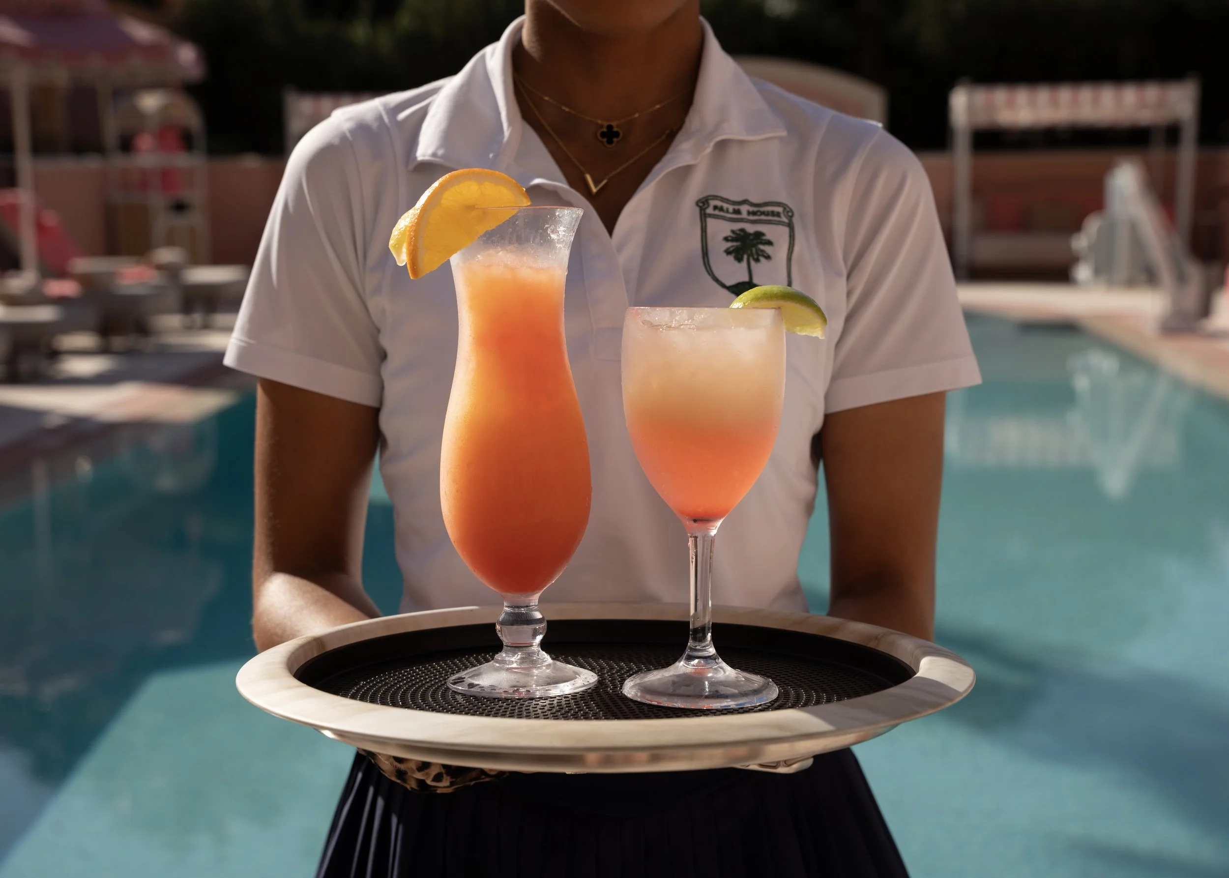 A waitress holds a tray with two tropical cocktails near a swimming pool, one garnished with a lemon wedge and the other with a lime wedge.