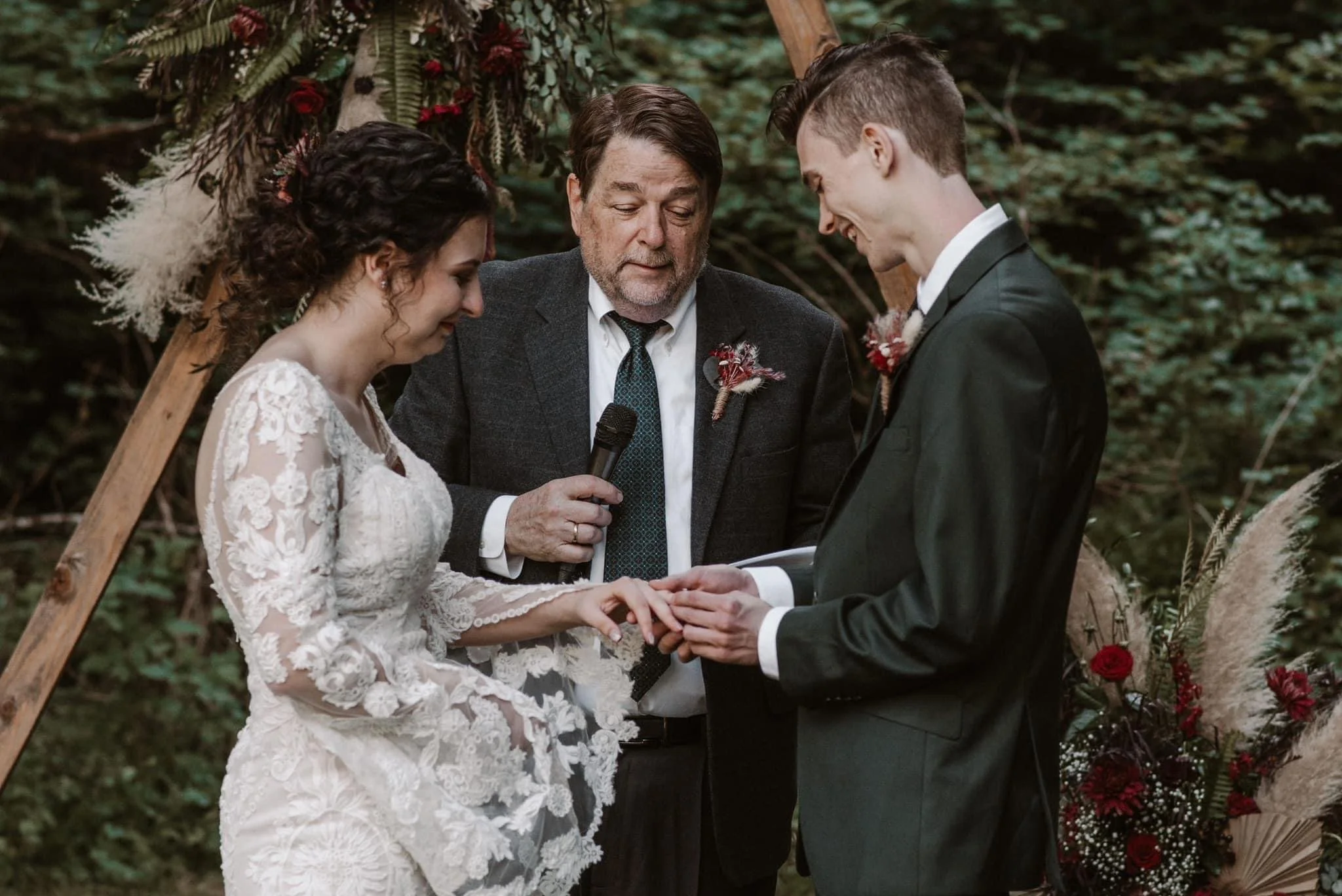 A couple getting married outdoors with an officiant, holding hands and exchanging rings, surrounded by flowers and greenery.