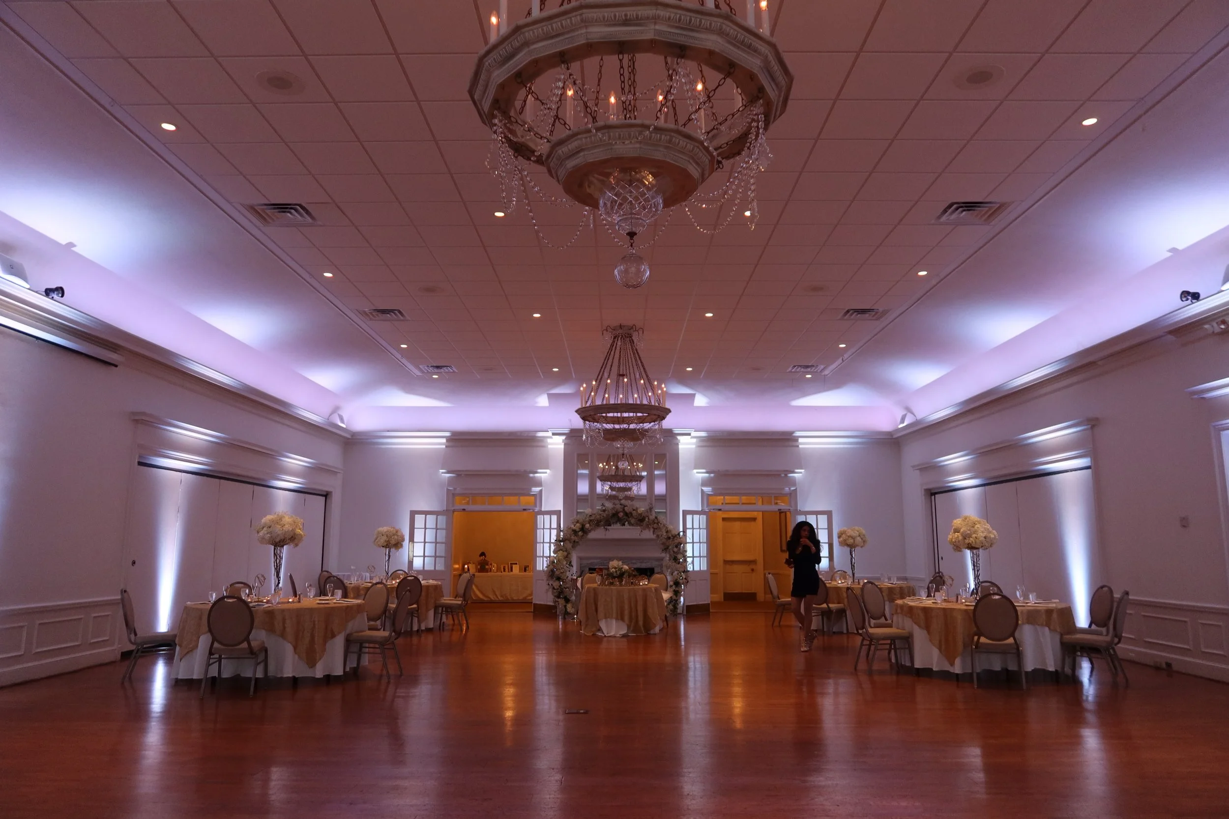 Elegant banquet hall with wooden floor, decorated for a wedding reception with a floral arch, round tables with white and gold tablecloths, floral centerpieces with white flowers, chandeliers, and wall sconces with purple lighting.