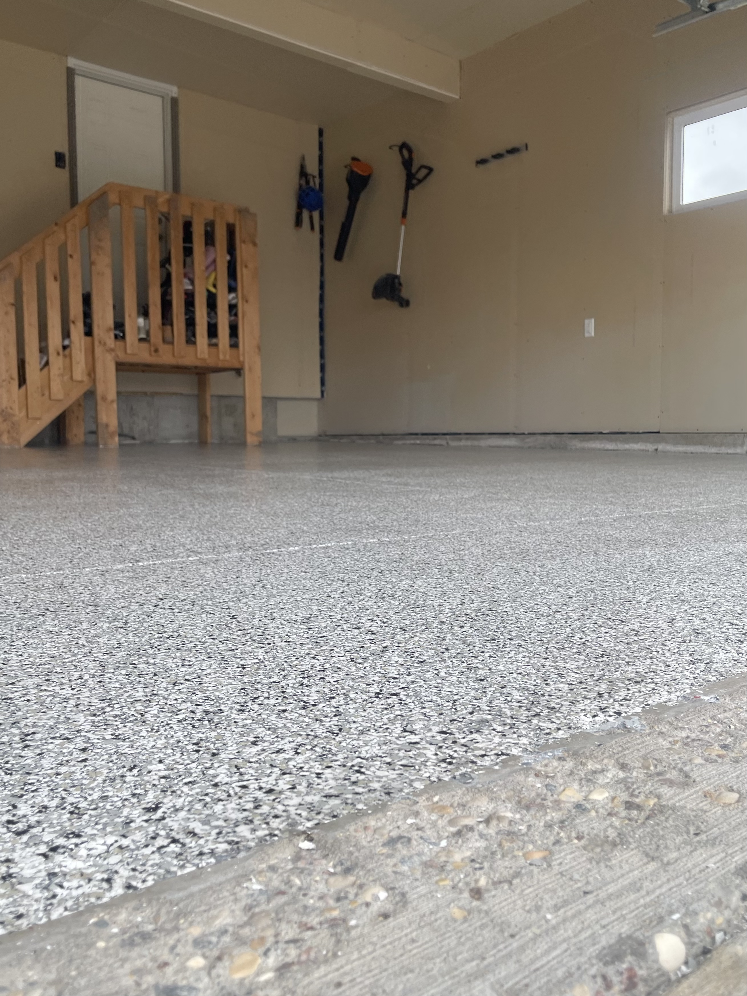 Empty garage with a speckled epoxy floor, wooden staircase, and tools hanging on the wall. Epoxy services in Winnipeg.
