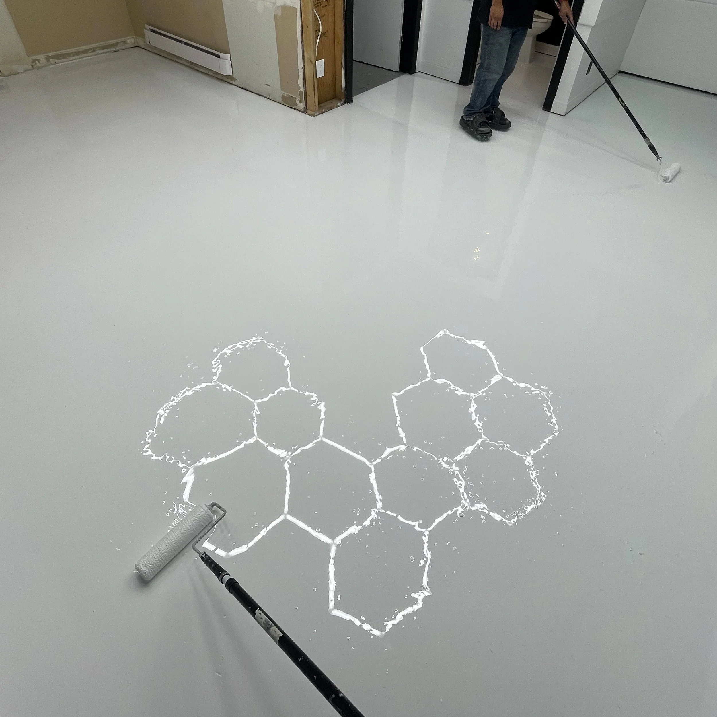 Person applying white epoxy resin on a floor with a roller, featuring a hexagonal pattern emerging from the coating. Epoxy services in Winnipeg.