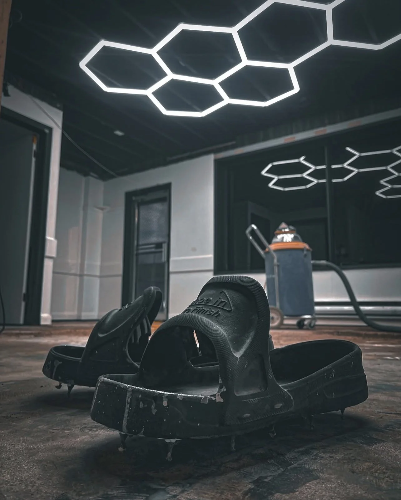 A pair of black ski boots on a platform in an indoor space with wooden flooring, a dust collector, windows, and a modern ceiling light fixture with hexagon-shaped lights.