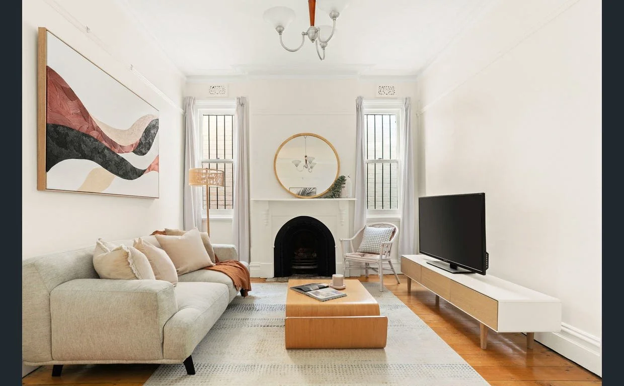 Modern living room with beige sofa, abstract artwork, coffee table, flat-screen TV on a stand, fireplace with a round mirror, and two windows with curtains.