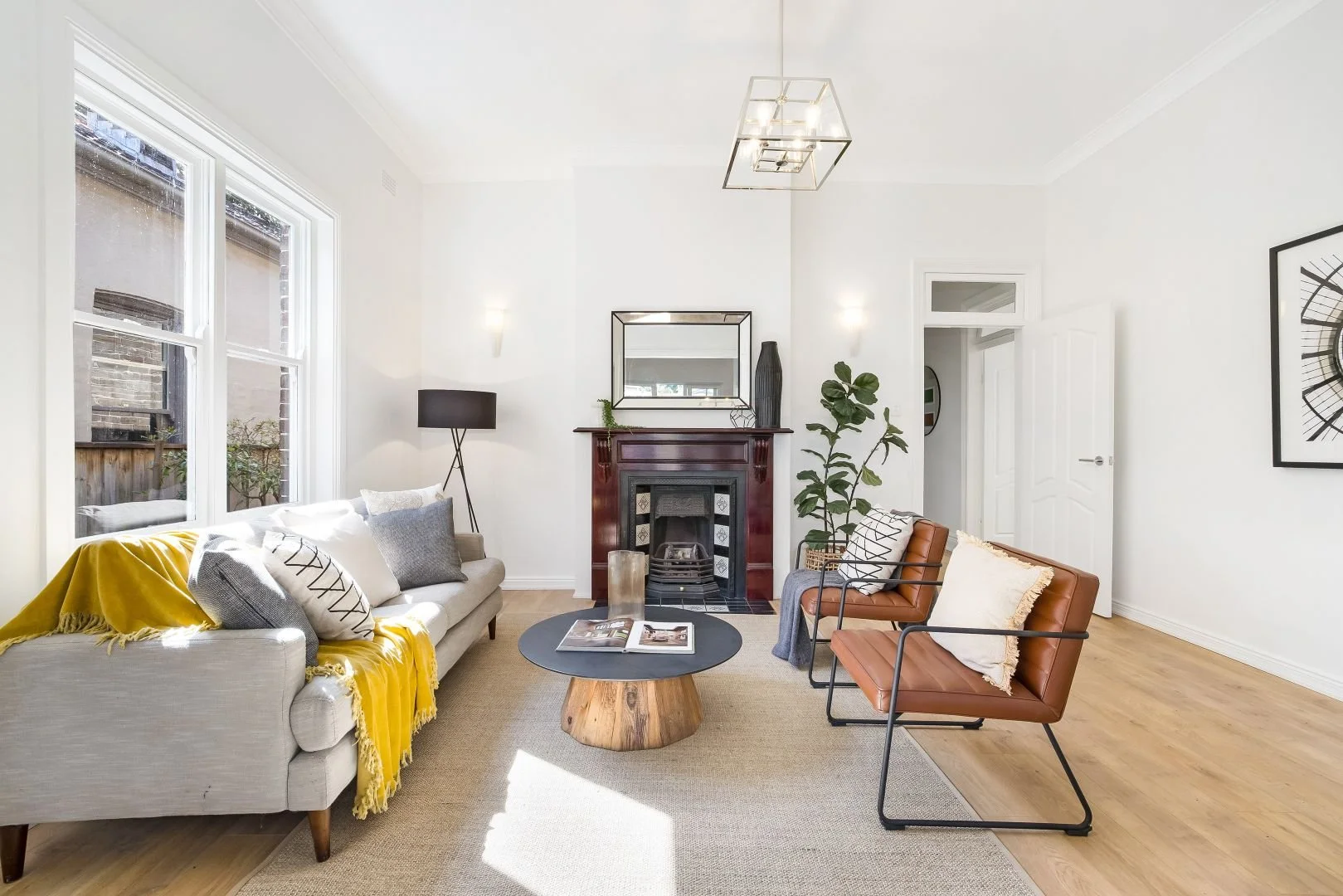 Modern living room with gray sofa, yellow throw, pillows, leather chairs, fireplace, coffee table, large window, floor lamp, indoor plant, and wall art.
