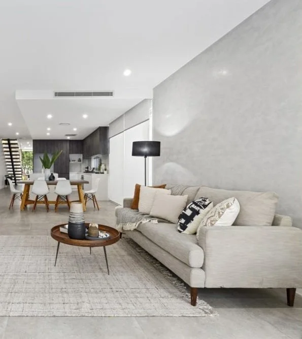 Modern living room with a beige sofa, decorative pillows, and a gray accent wall. A round wooden coffee table with vases is on a light patterned rug. In the background, there's a dining area with a wooden table, white chairs, and a kitchen with dark cabinets. A floor lamp and greenery add to the decor.