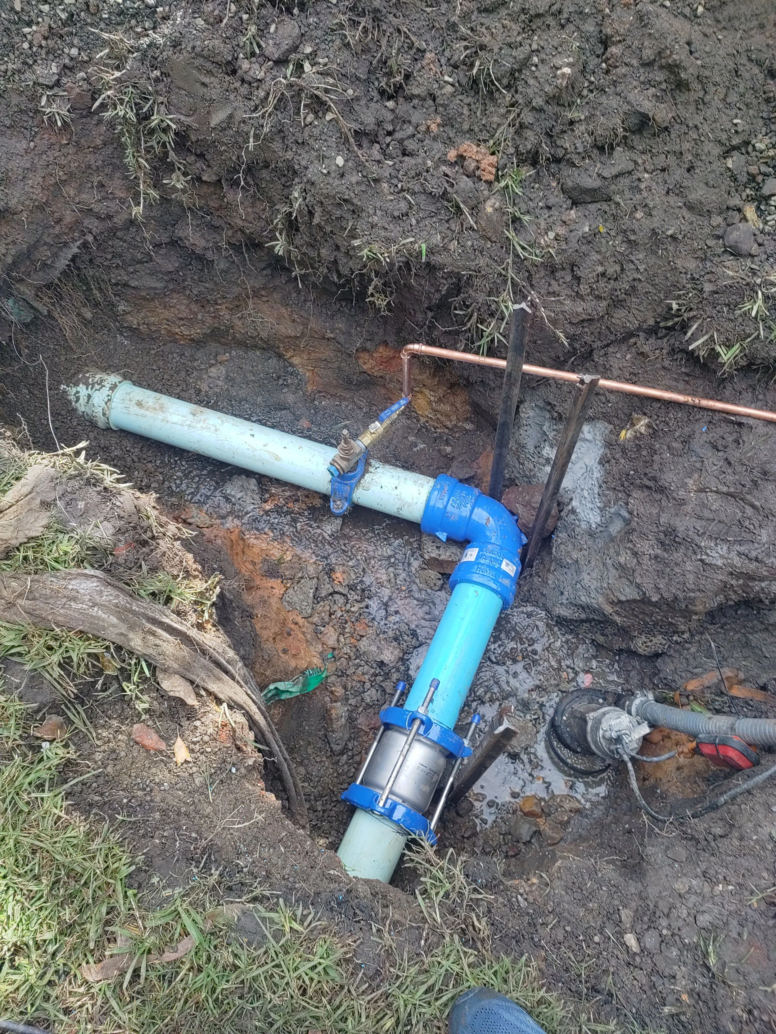 Exposed underground plumbing with blue PVC and metal pipes, connectors, and valves in a muddy pit.