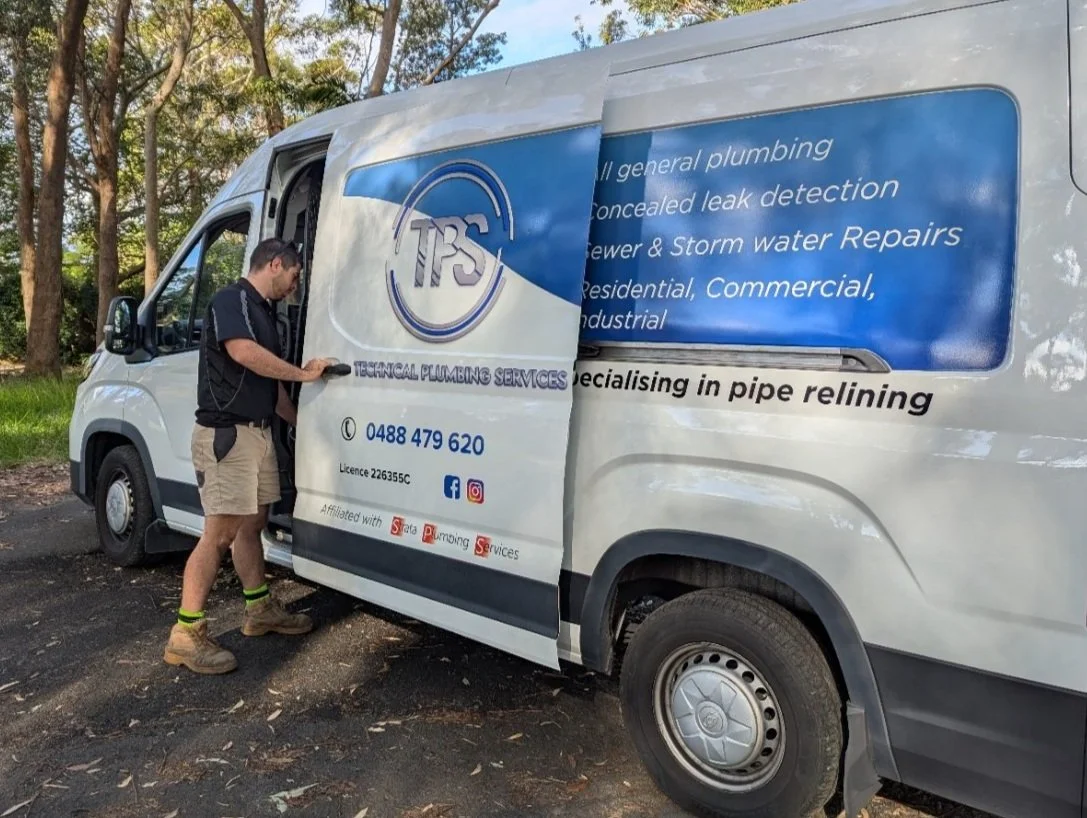 Man opening the sliding door of a plumbing service van in a wooded area, with company signage and contact information displayed on the van's side.