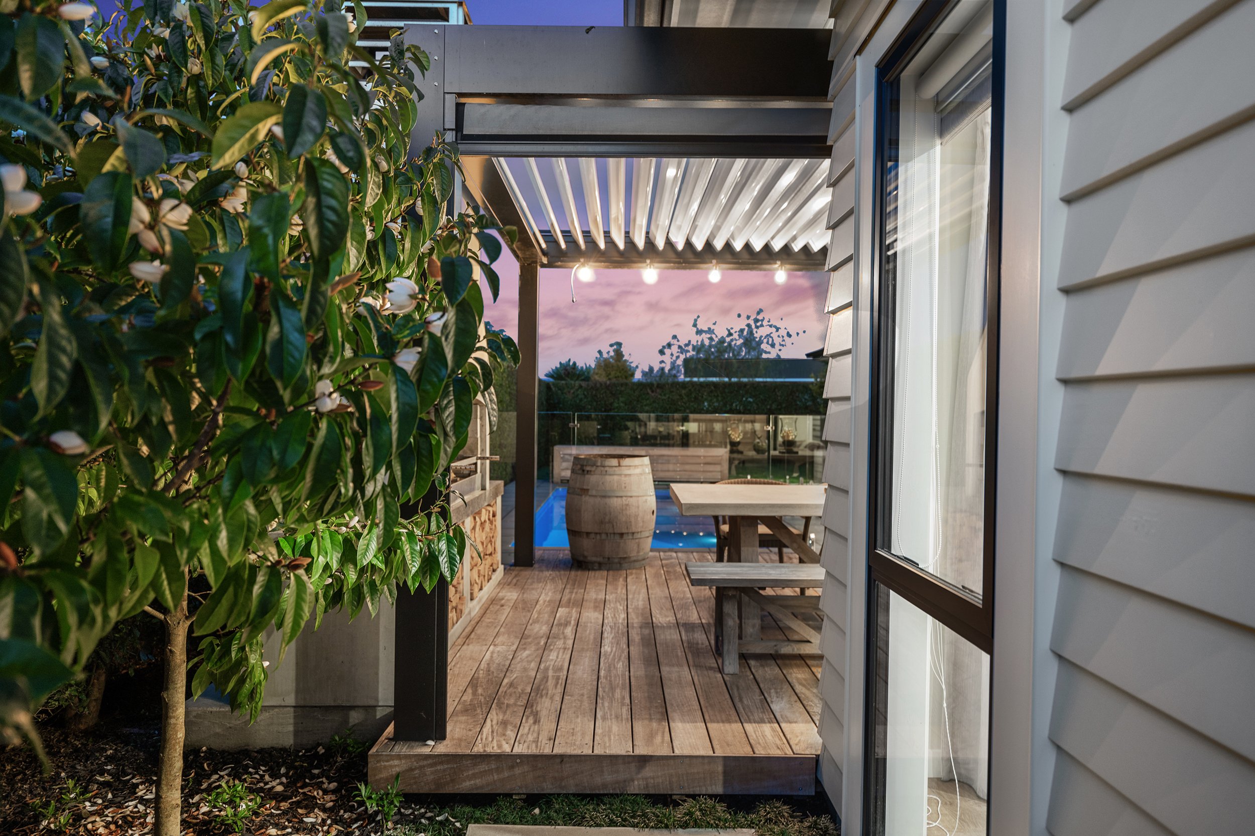 Outdoor wooden deck with pergola, string lights, a wooden table with benches, a large barrel, and a nearby tree with white flowers. A pool and glass fence are in the background.