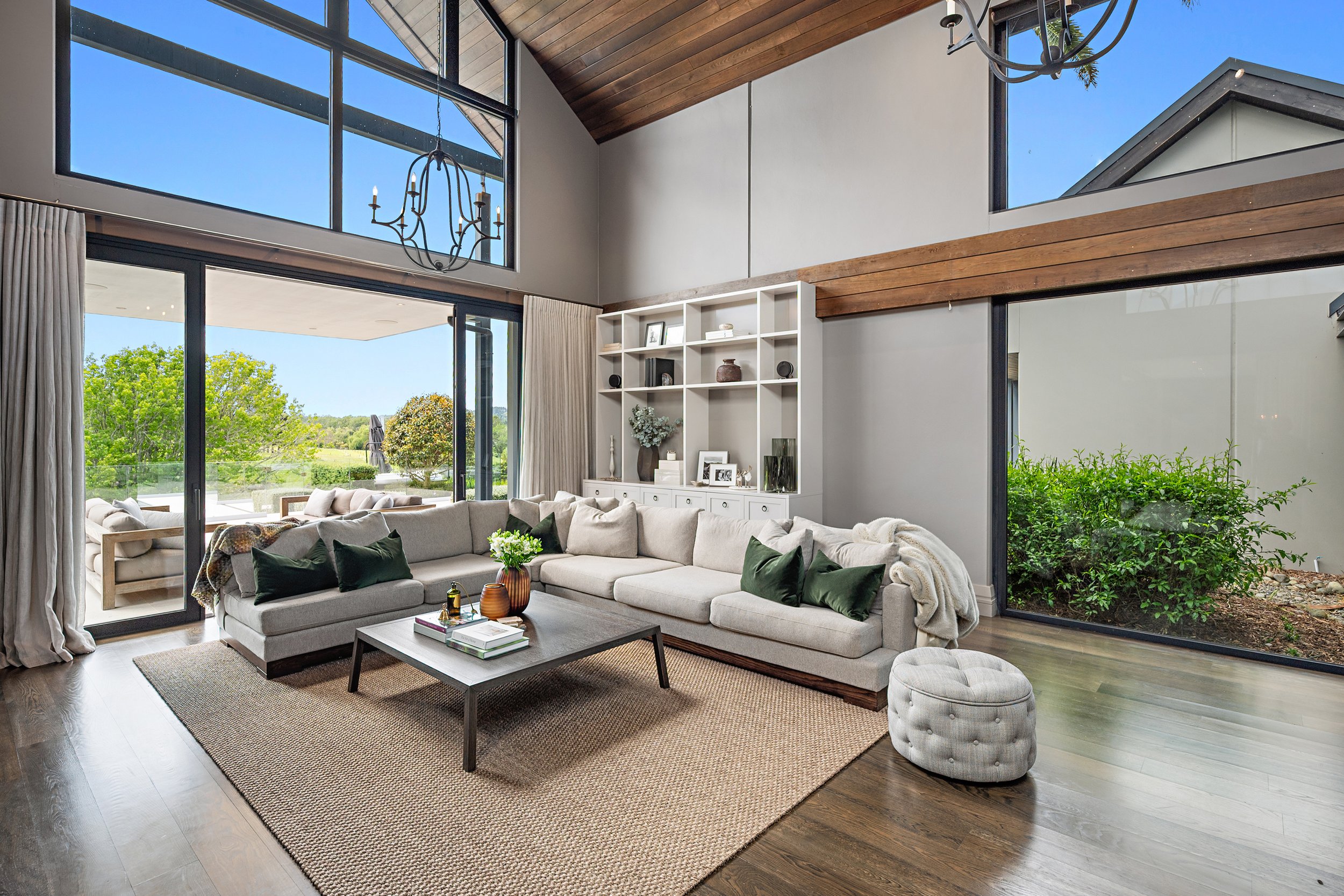 Spacious modern living room with high ceilings, large sectional sofa, hardwood floors, and floor-to-ceiling windows showcasing outdoor greenery.