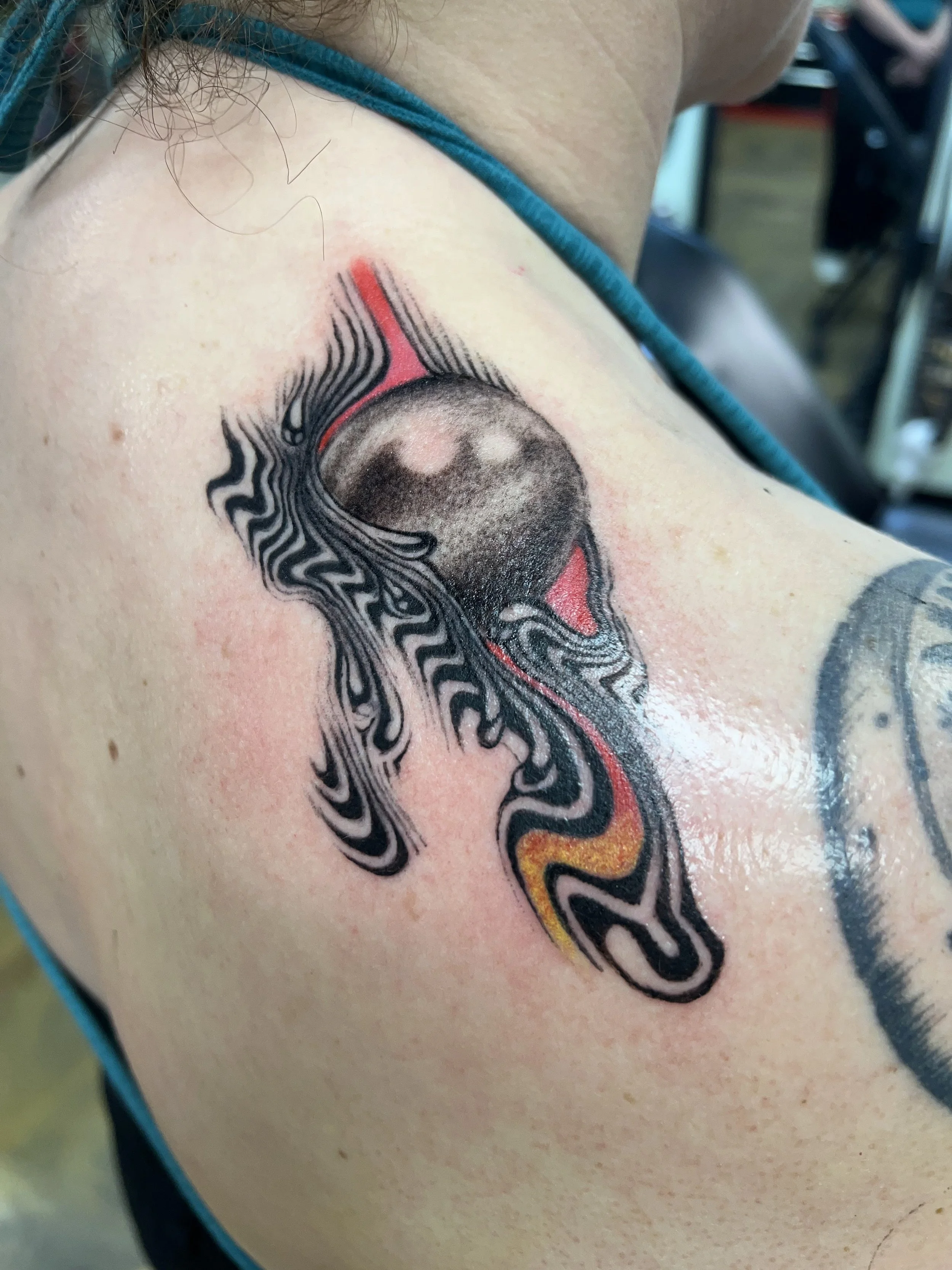 Close-up of a colorful tattoo on a person's shoulder featuring a stylized eye with black and red ink, surrounding abstract designs and lines.