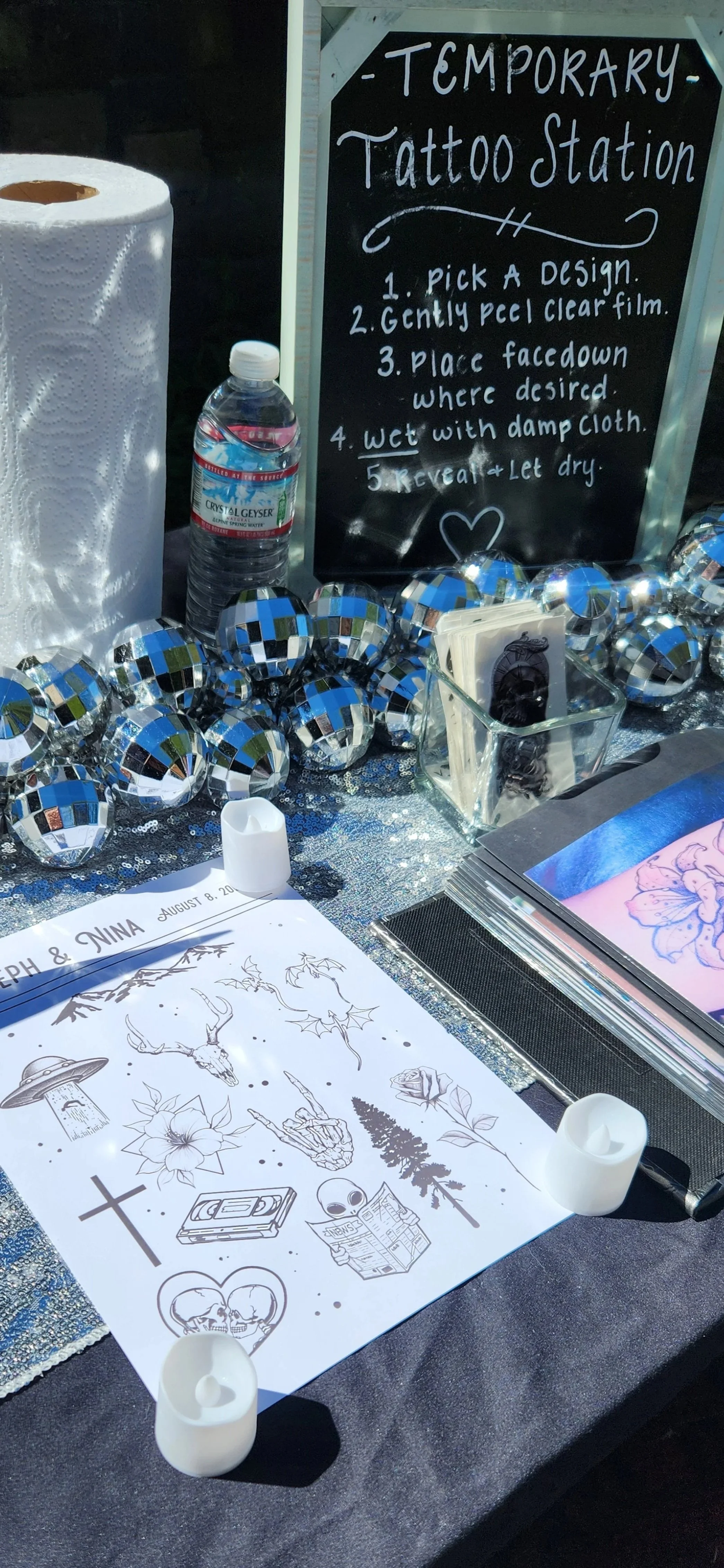 A sign for a temporary tattoo station with instructions, a bottle of water, and decorative blue, white, and black reflective ornaments on a table with tattoo designs and related materials.