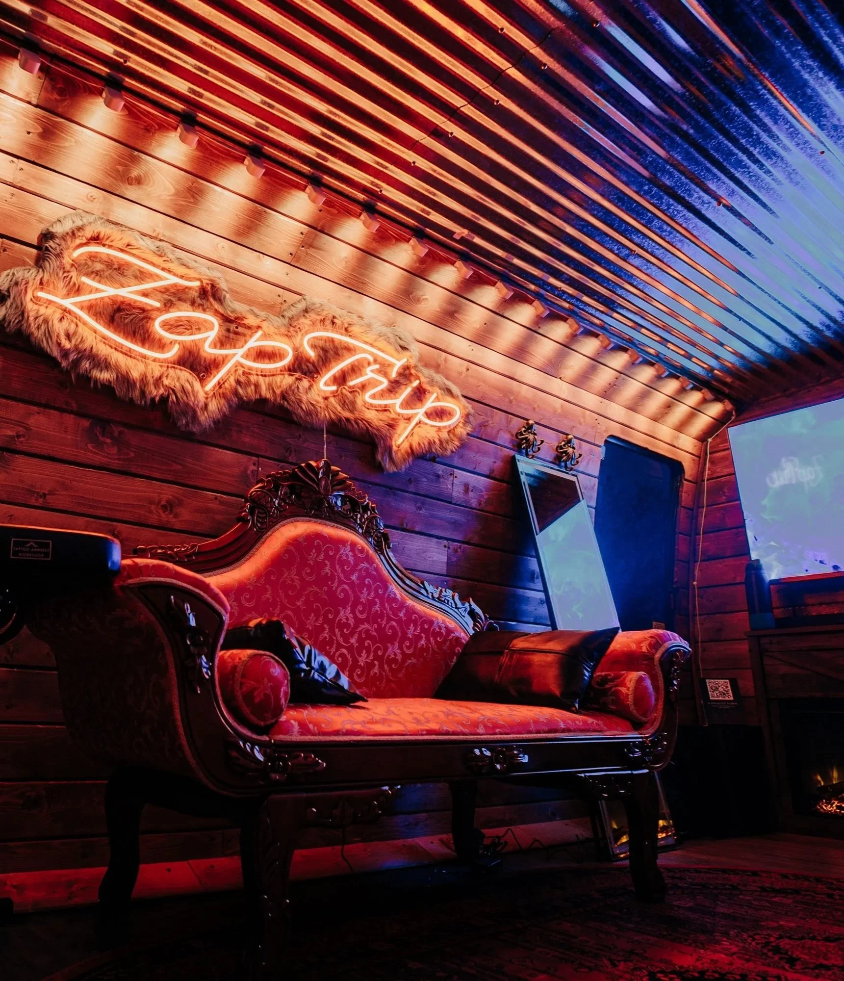 A cozy room with wooden walls and ceiling, featuring a neon sign that says 'Happy' with fur trim, a vintage ornate sofa with red upholstery, and a mirror and TV.