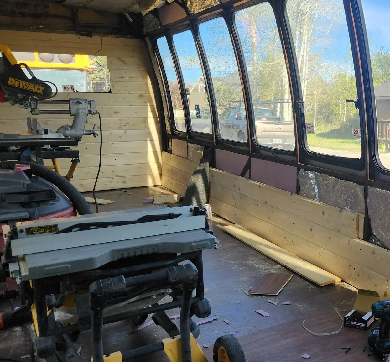 Interior of a room under renovation with wooden wall paneling on the right side, construction tools including a miter saw and a vacuum cleaner, and large windows letting in natural light, revealing a street and parked vehicles outside.